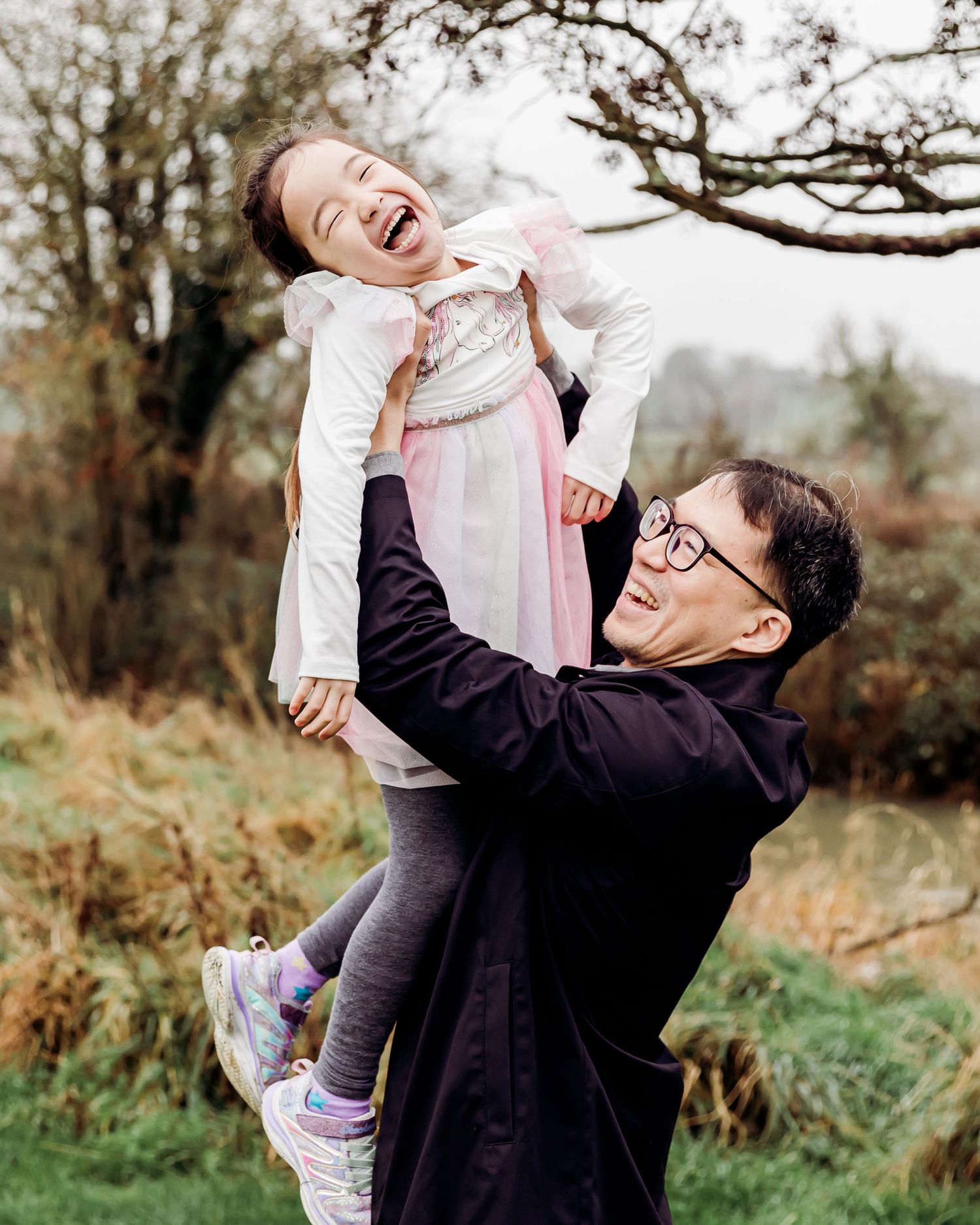 A joyful father lifts his laughing young daughter outdoors in a lush green park setting.