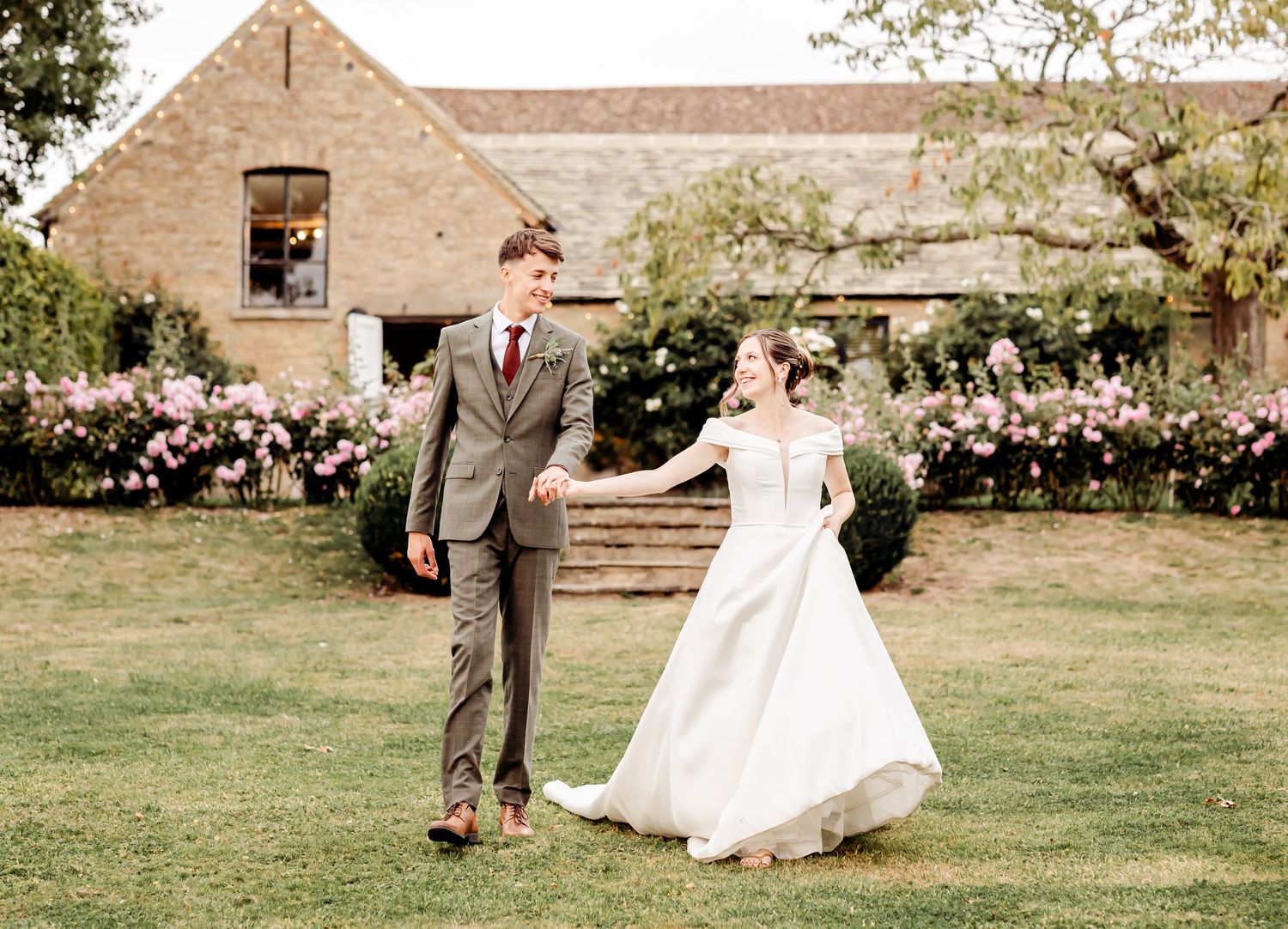 Bride and groom holding hands walking through garden with blooming roses and barn backdrop.