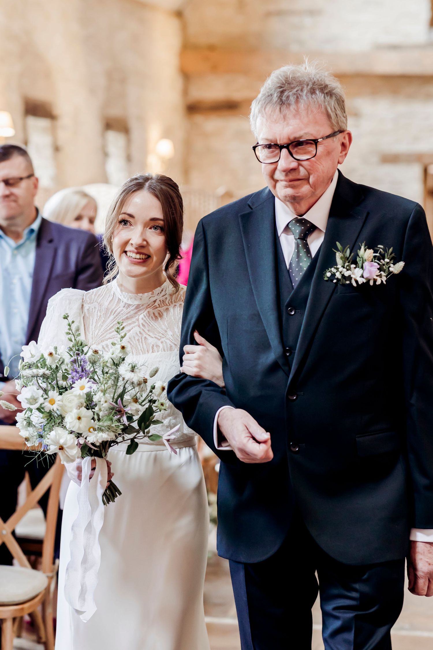 Bride in lace wedding dress holding wildflower bouquet walks down aisle with older man in dark suit.