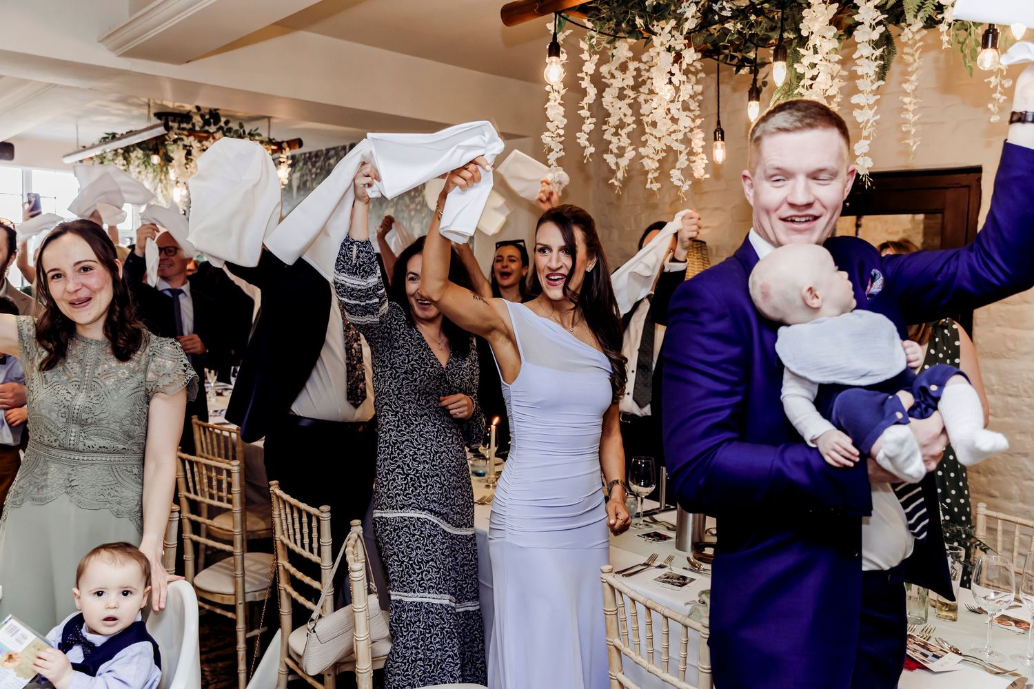 Joyful wedding guests wave napkins and celebrate at lively indoor wedding reception.