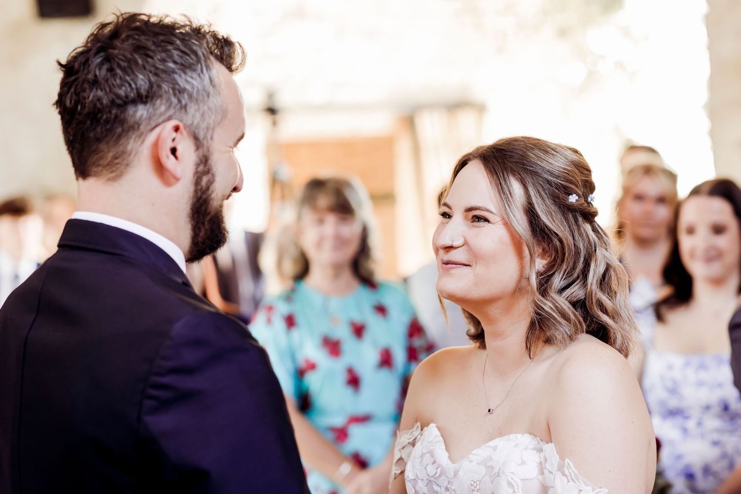 Bride gazes lovingly at groom during wedding ceremony with guests in background.