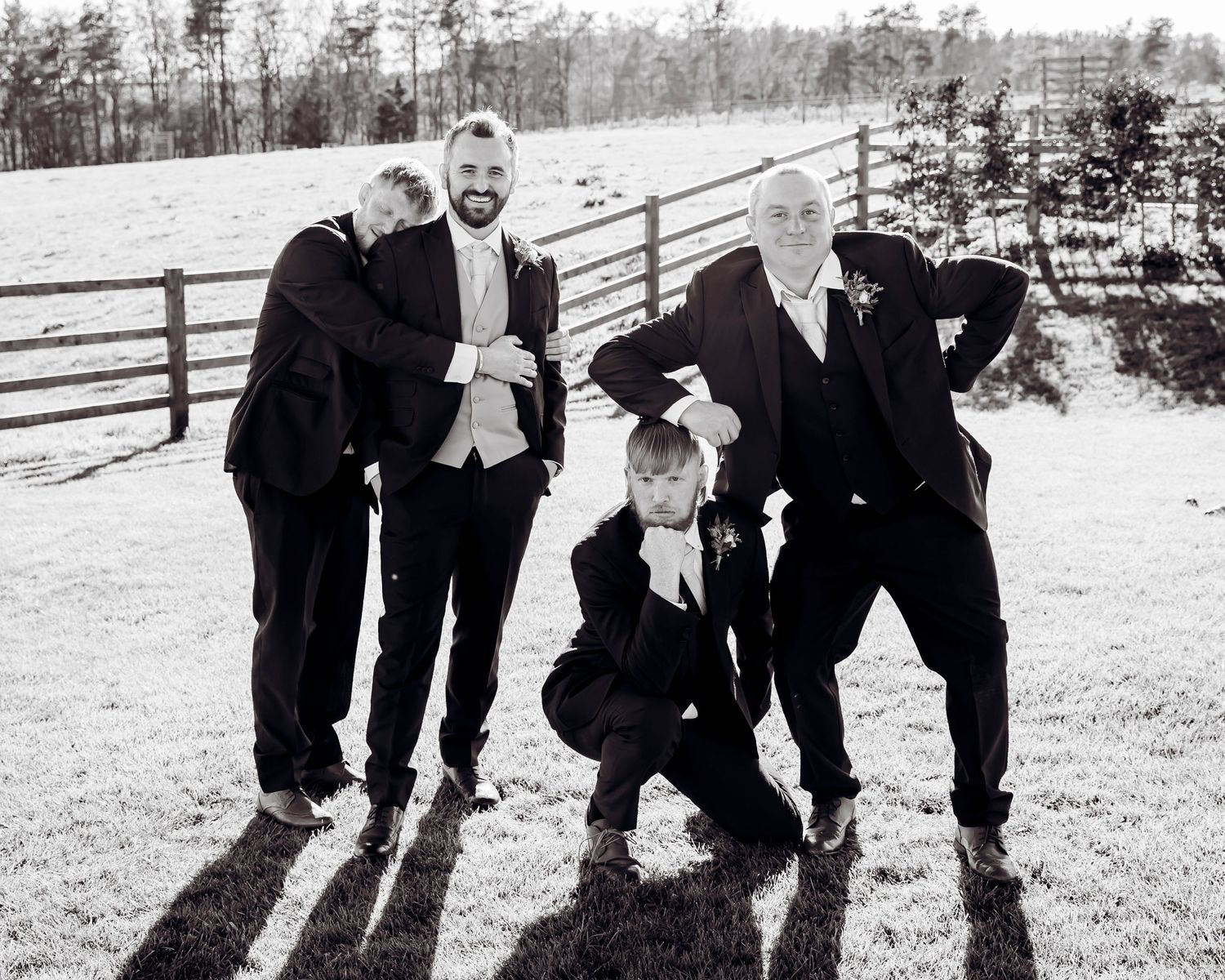 Black and white photo of groom posing playfully with groomsmen and young boy in suits at outdoor farm venue.
