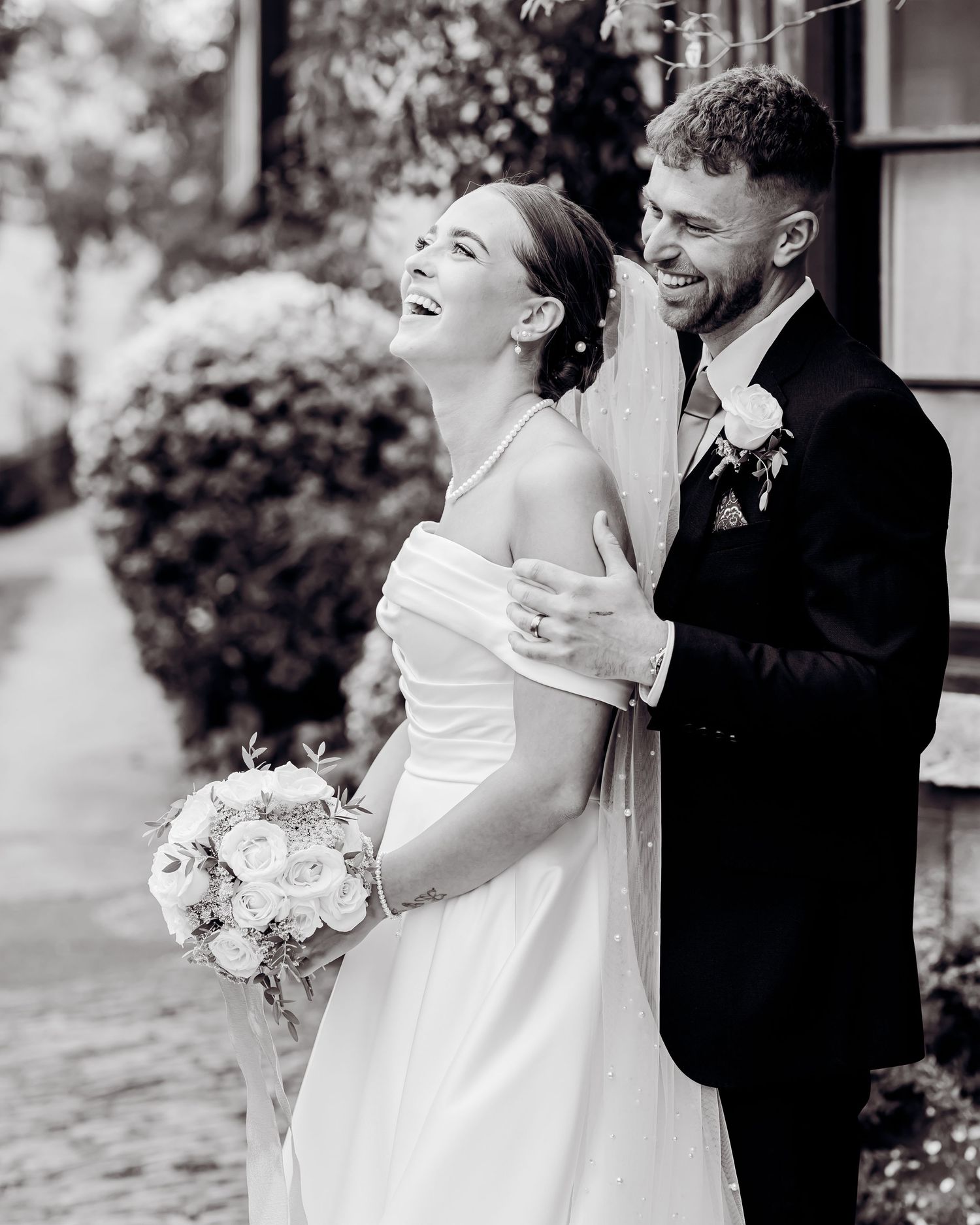 Black and white photo of laughing bride holding bouquet embraced by groom outdoors.