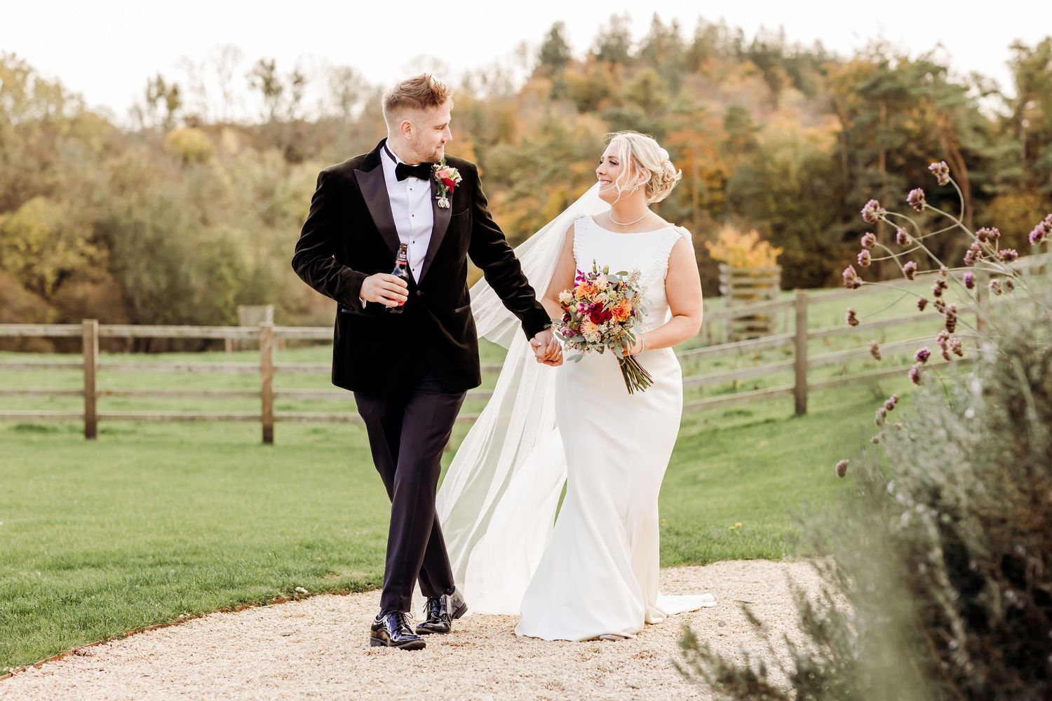 Bride and groom walking hand in hand through scenic countryside path with autumn foliage in background.