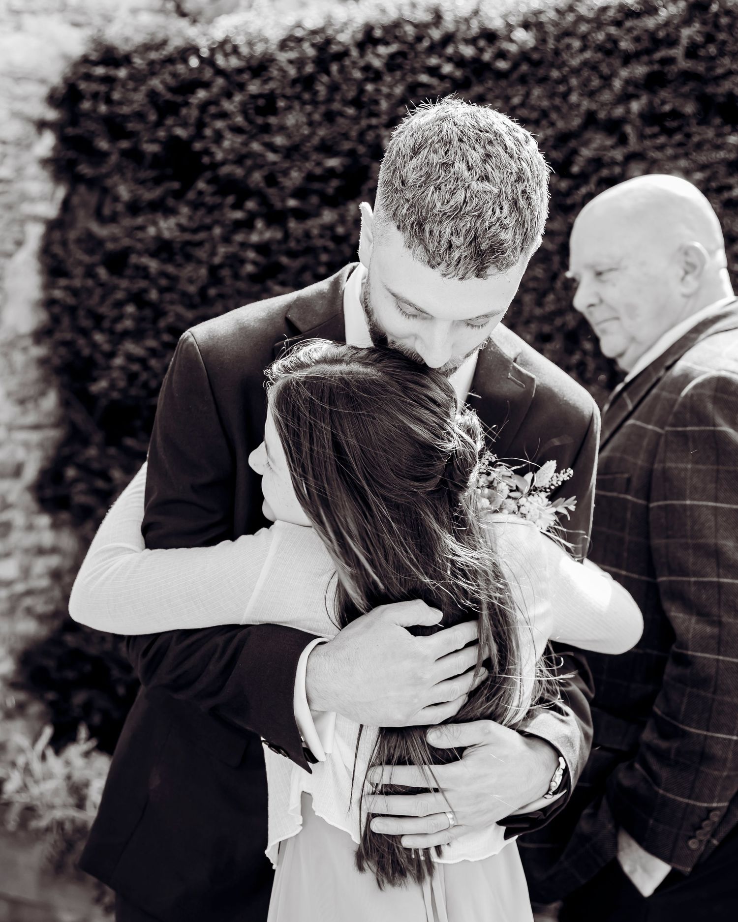 Emotional black and white wedding photo of groom embracing bride with guest watching in background.