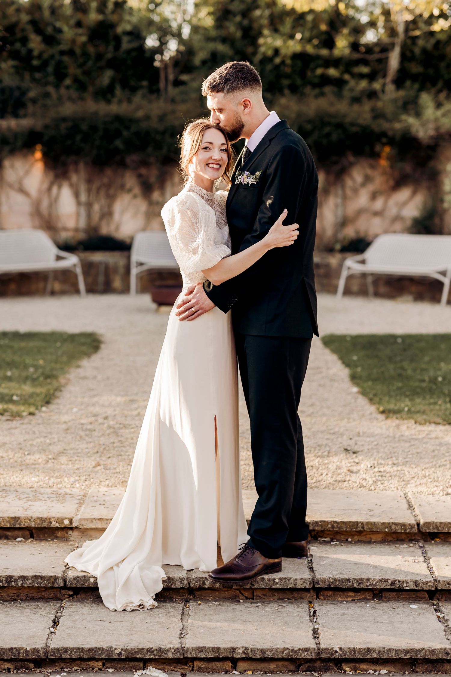 Bride and groom embracing outdoors in golden hour light, bride in white gown, groom in black suit.