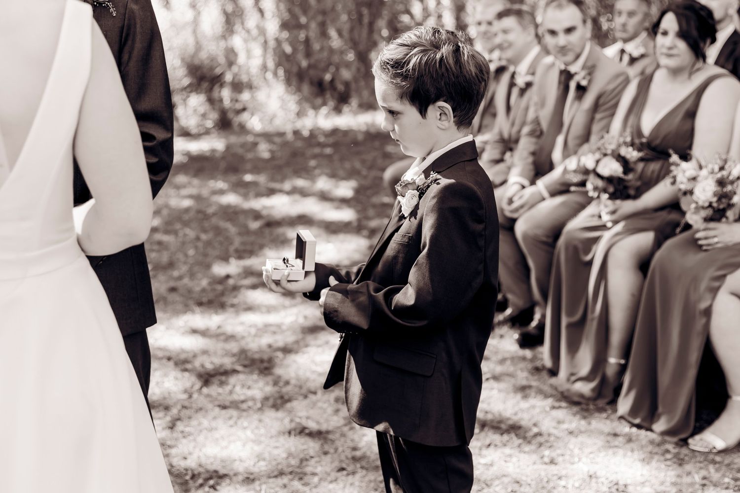 Young ring bearer in black suit presenting rings during an outdoor wedding ceremony with guests seated in background.