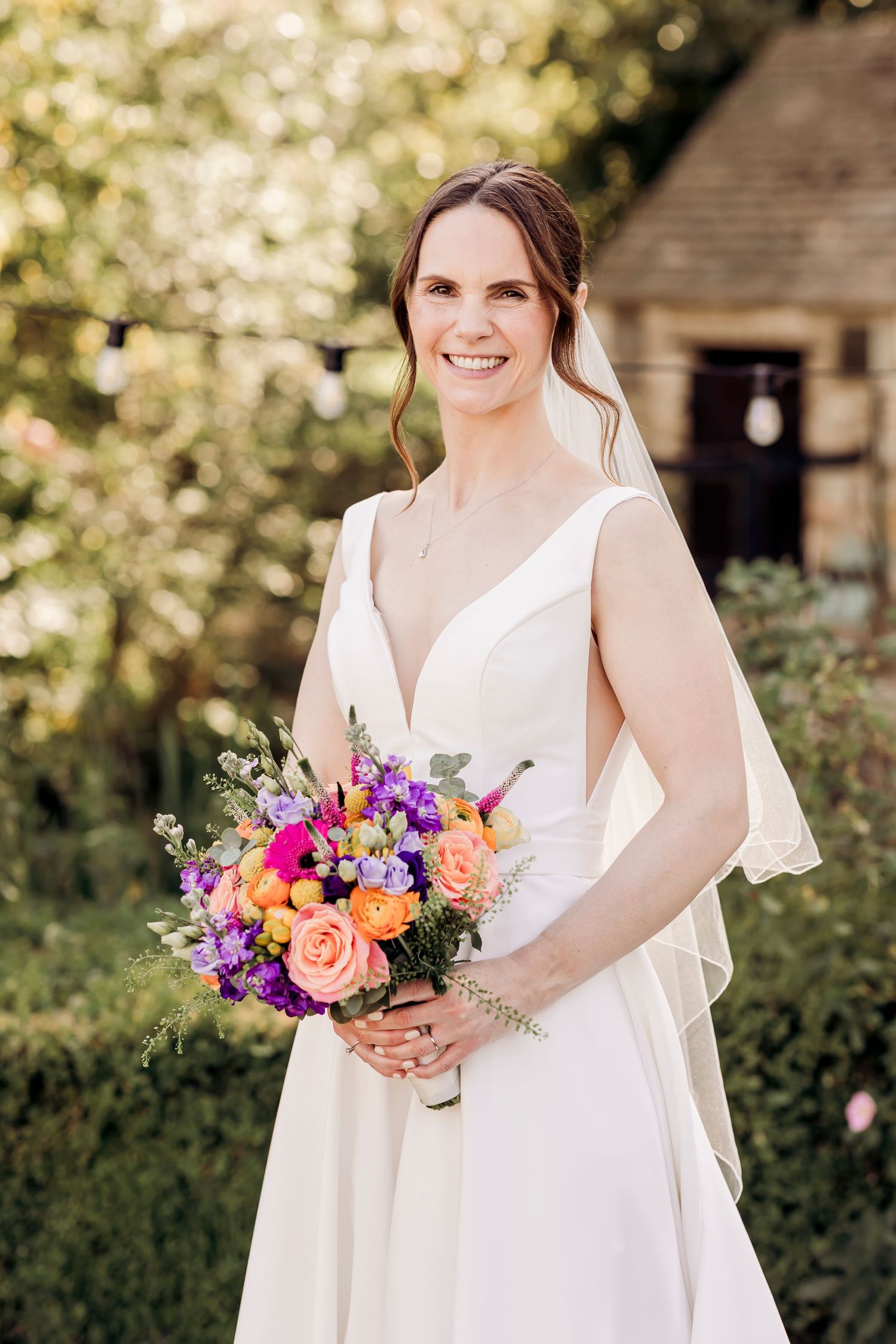 Smiling bride in white V-neck gown and veil holding vibrant orange and purple bouquet outdoors.