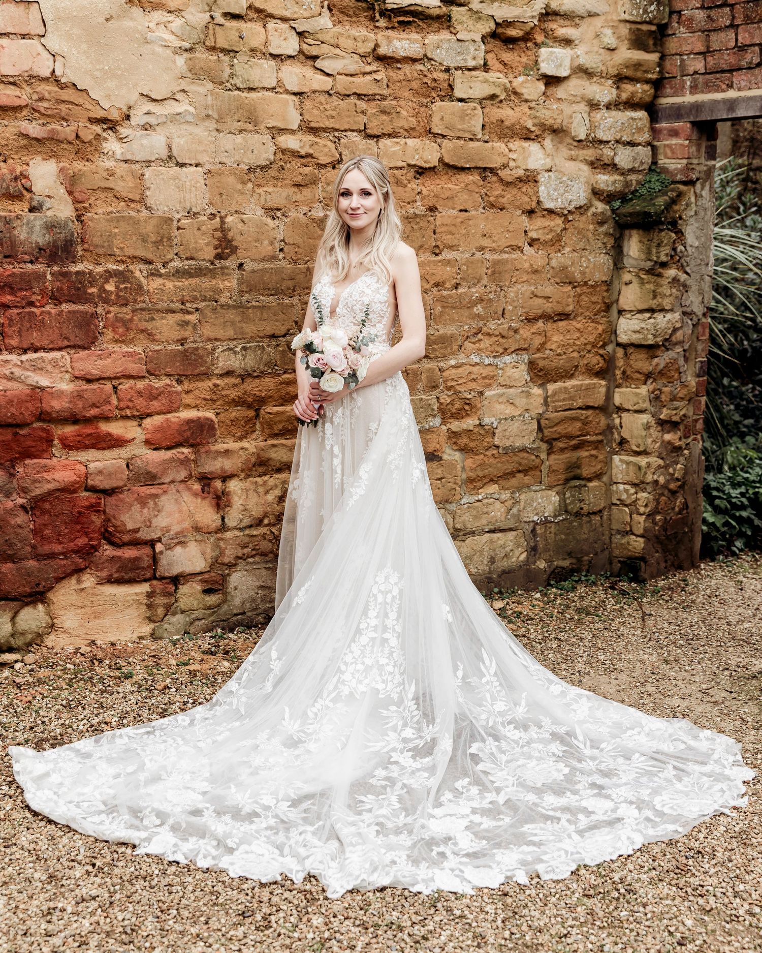 Bride in a stunning lace A-line gown with long train holding bouquet against rustic brick wall.