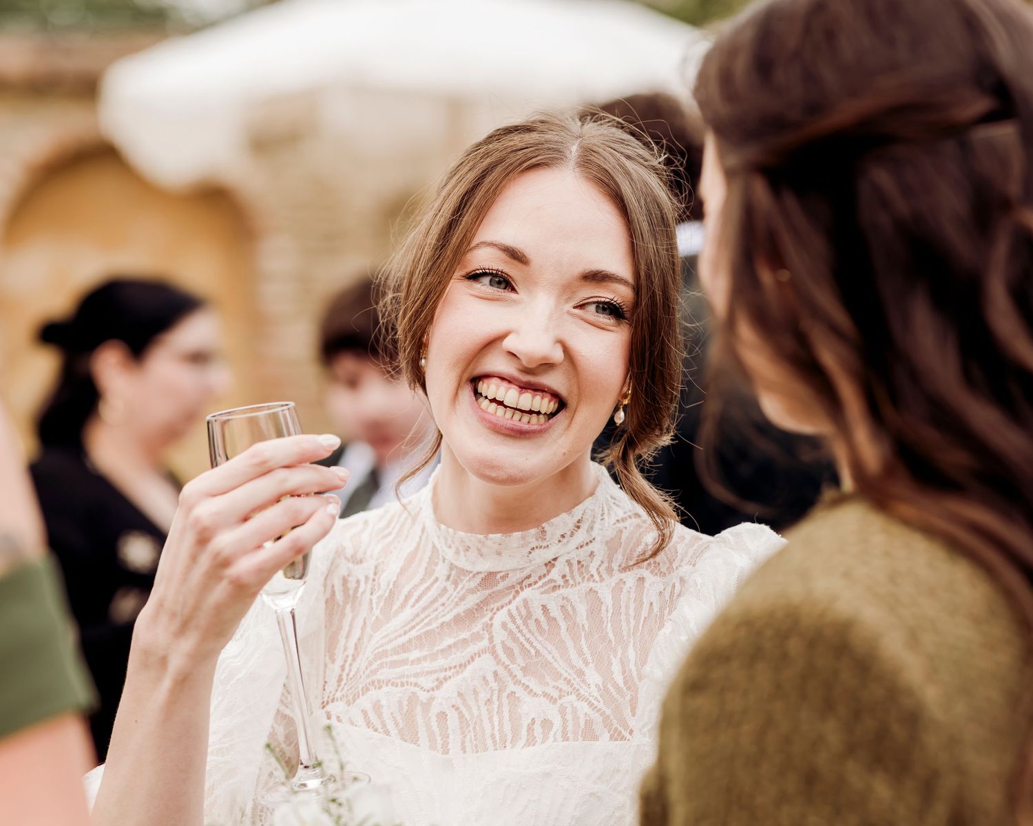 Joyful bride in white lace dress laughing while holding a drink at her wedding reception.