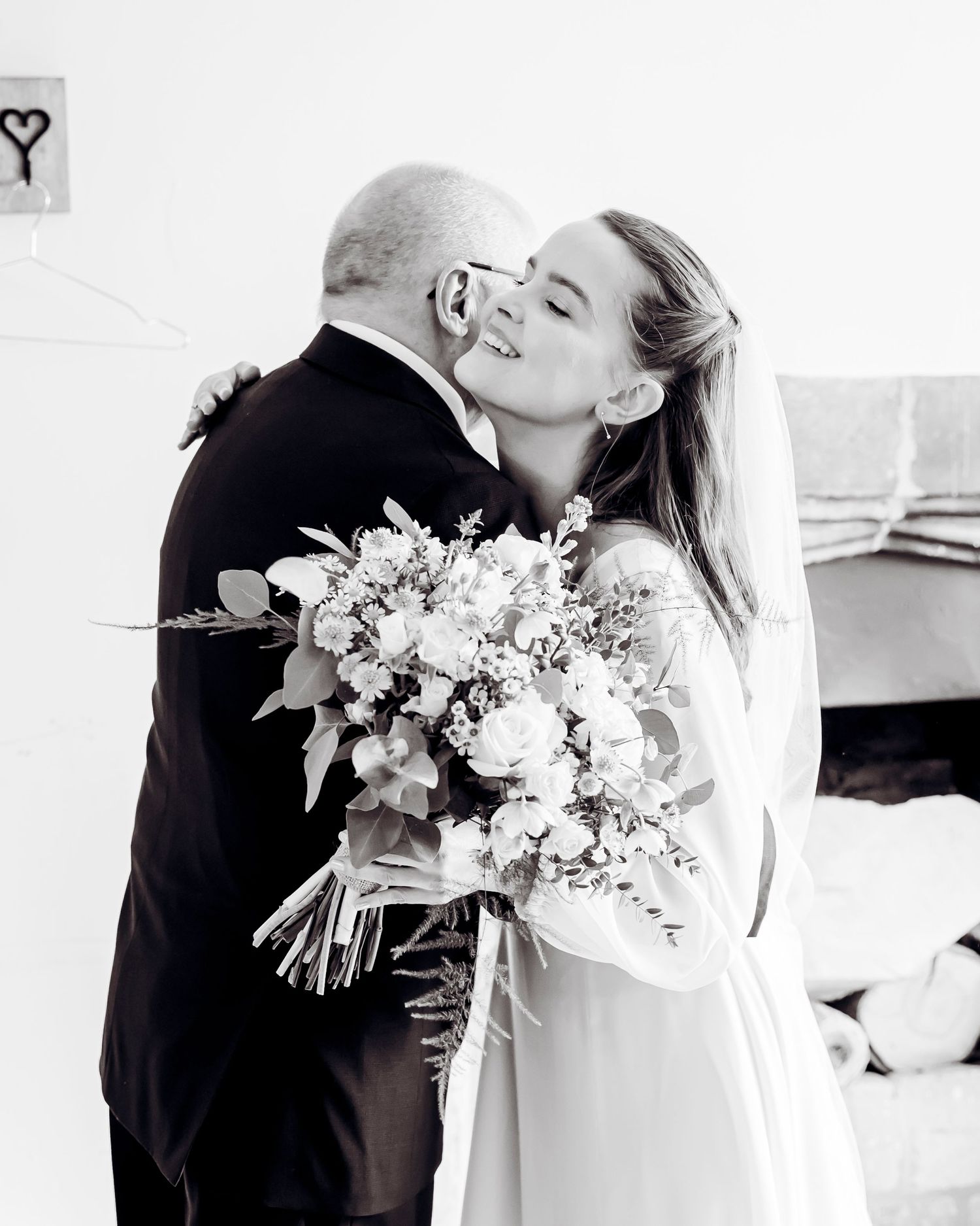 Smiling bride in white dress holding a lush bouquet receiving a kiss from her father in black and white.