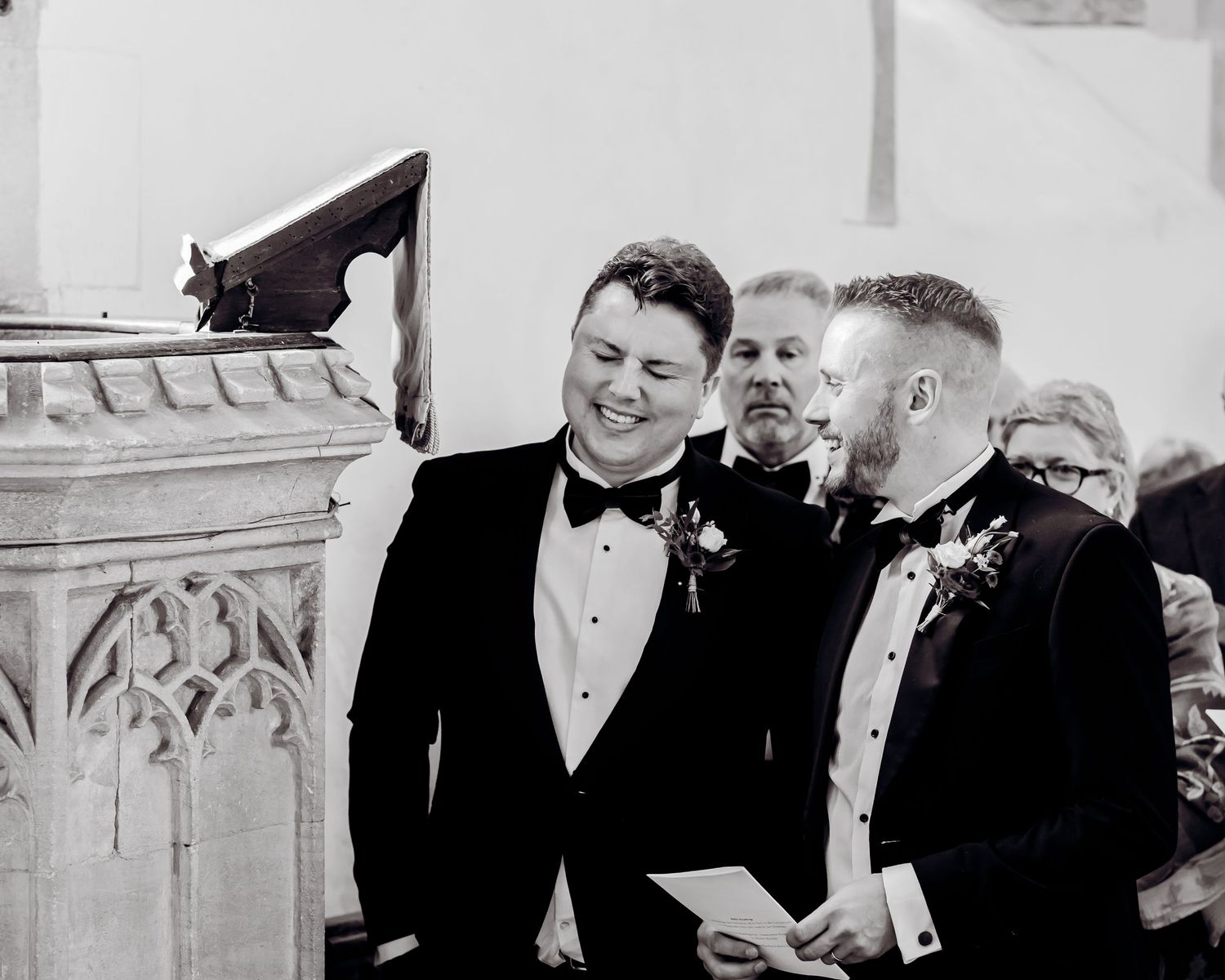 Two groomsmen laughing together in black tuxedos during a wedding ceremony in black and white photo.