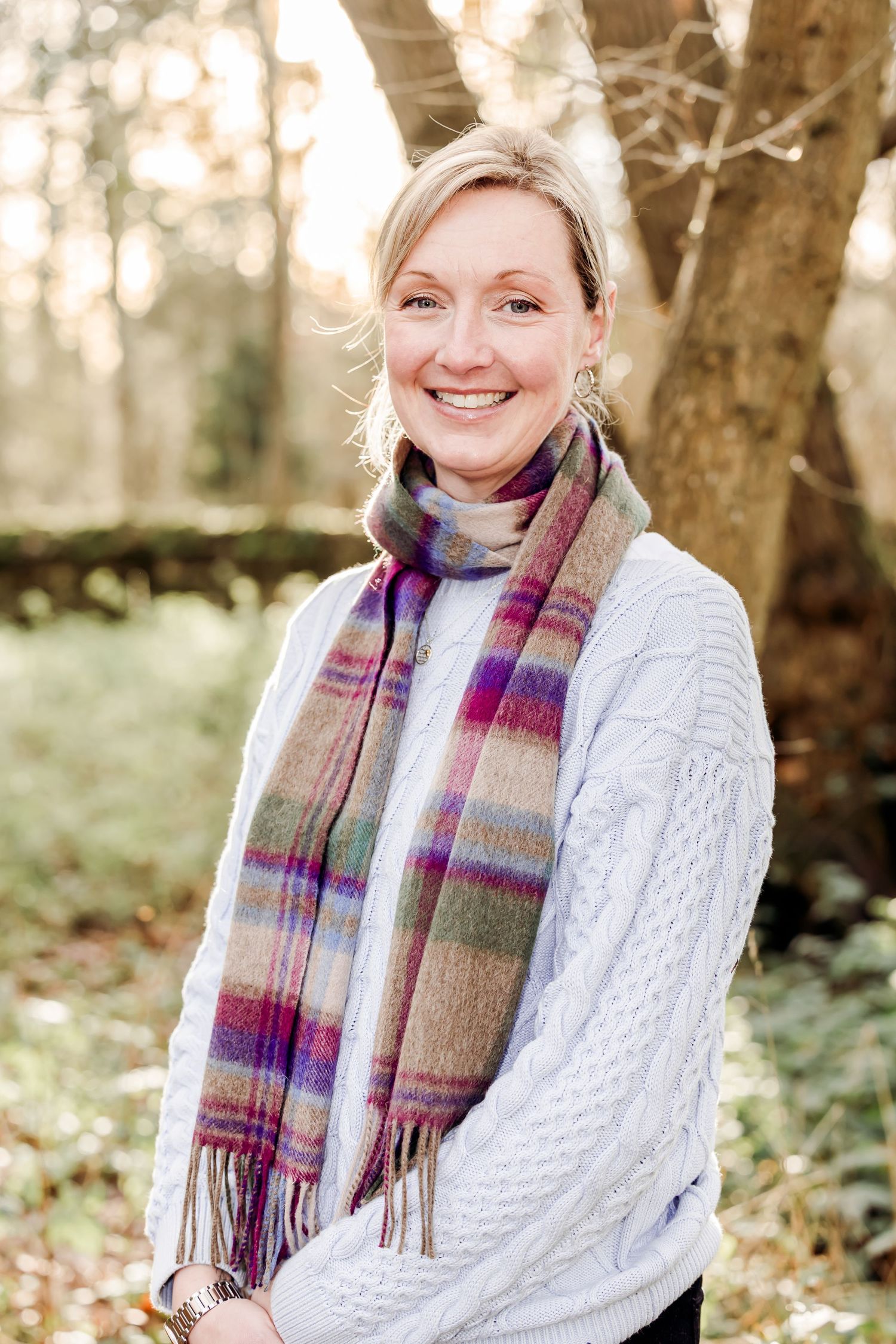 Cheerful blonde woman in white cable knit sweater and multicolored plaid scarf posing in autumn woodland.