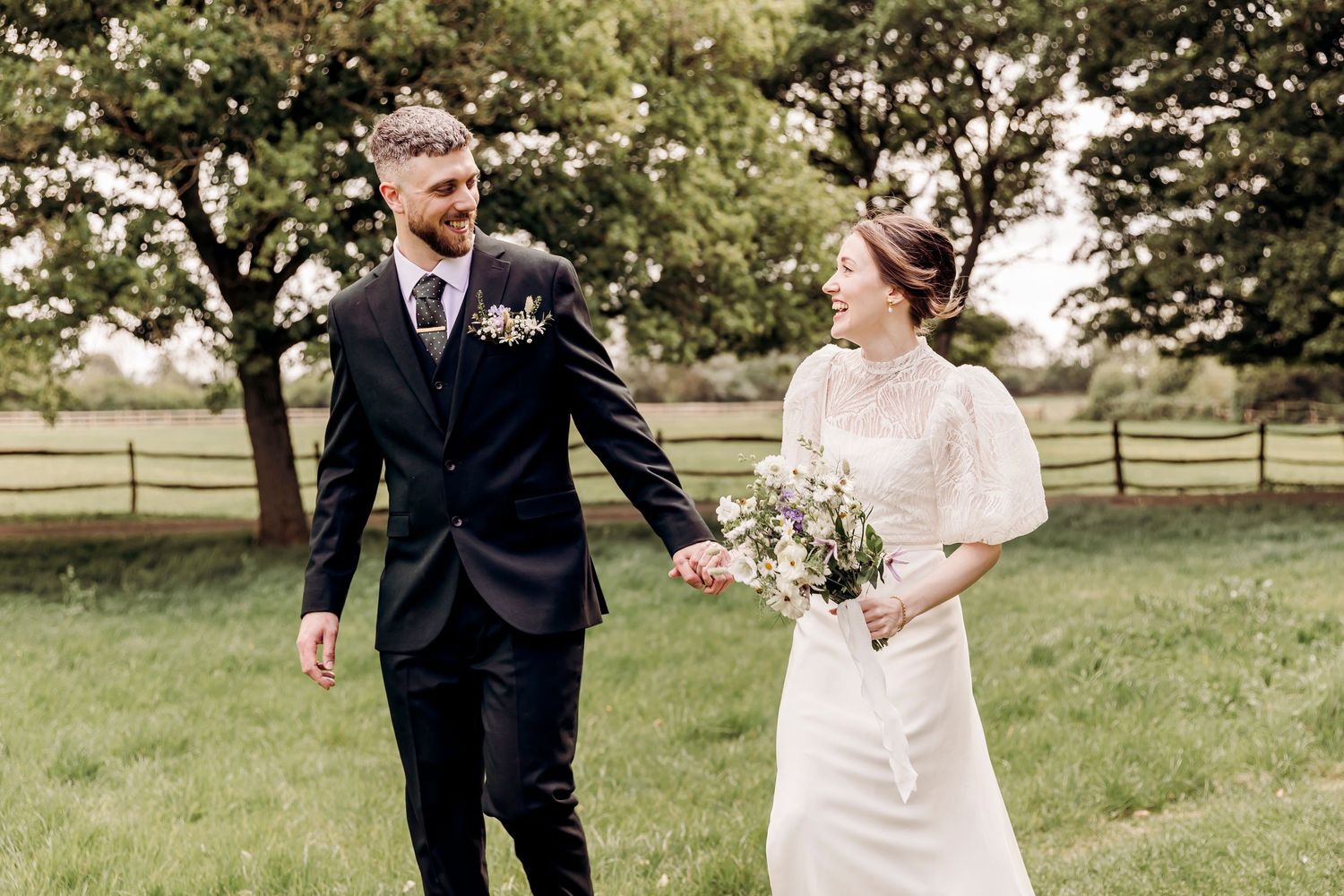 Happy bride and groom walk hand in hand through a green meadow on their wedding day.