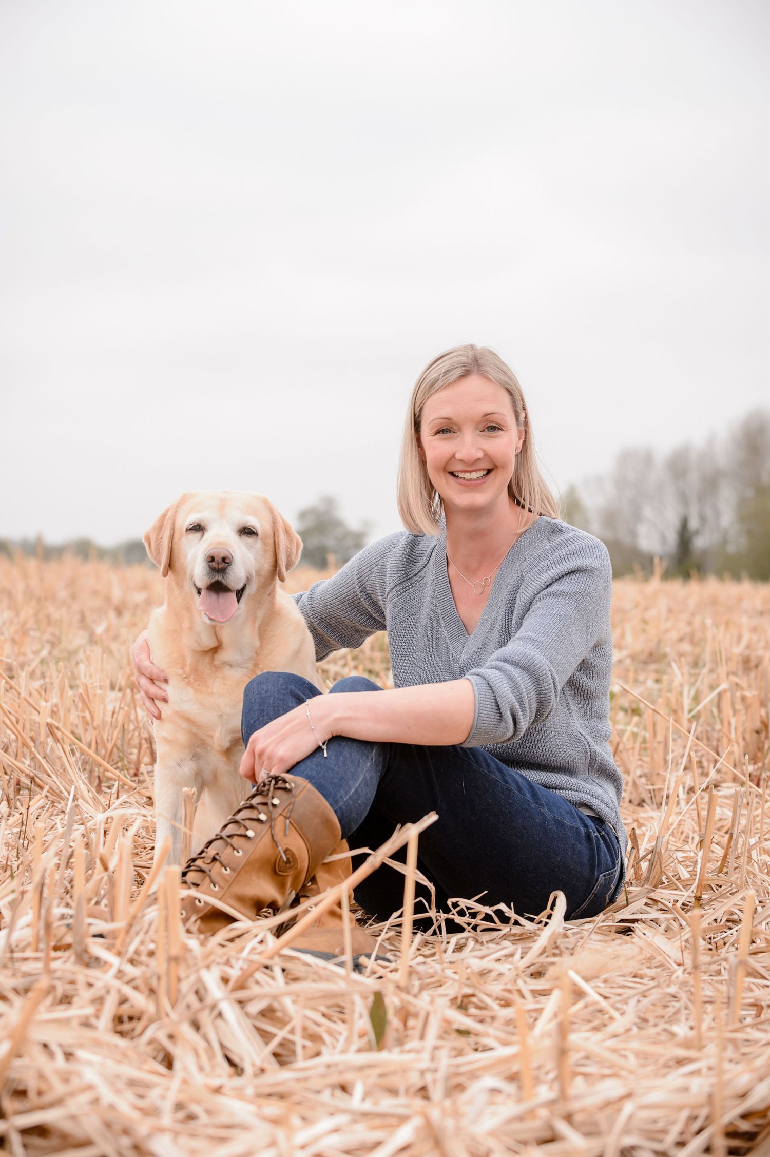 Smiling blonde woman sitting in a harvested field with a yellow Labrador Retriever dog on a cloudy day.