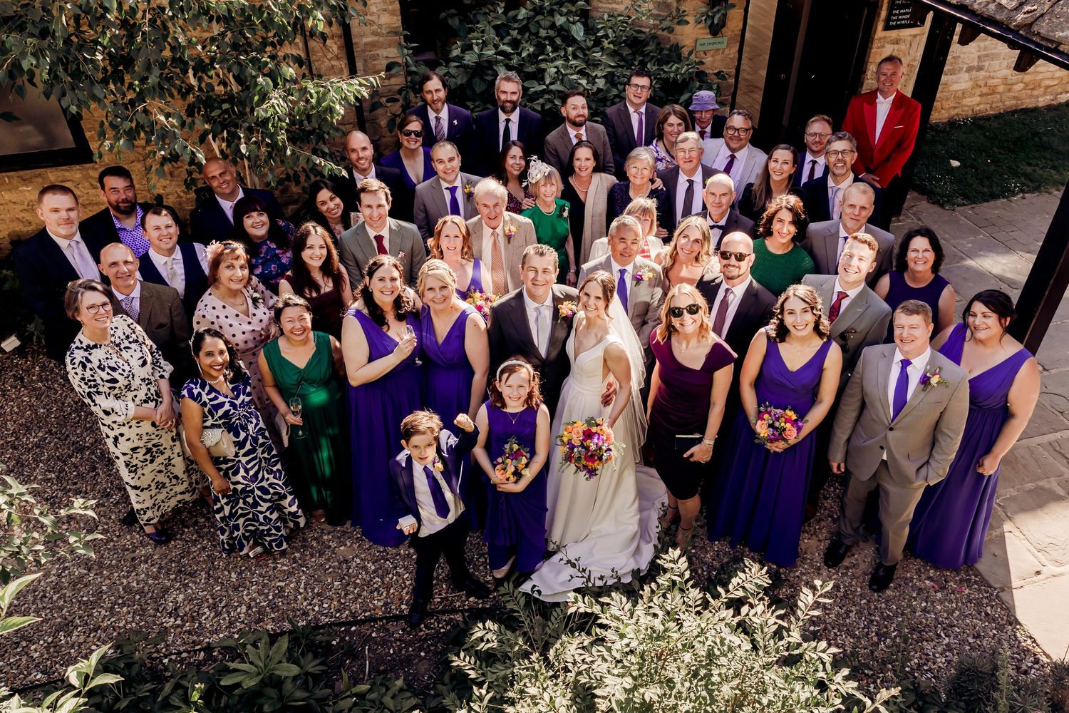 Large wedding party group photo with bridesmaids in purple dresses and guests gathered outdoors.