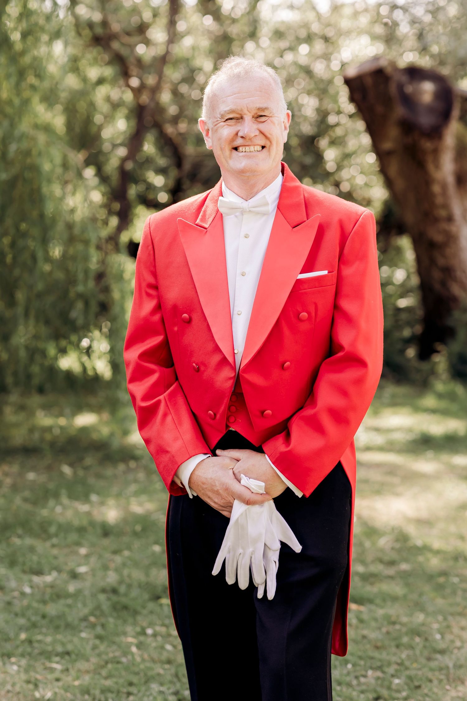 Smiling man in red tailcoat jacket with white gloves and bow tie posing outdoors at formal event.