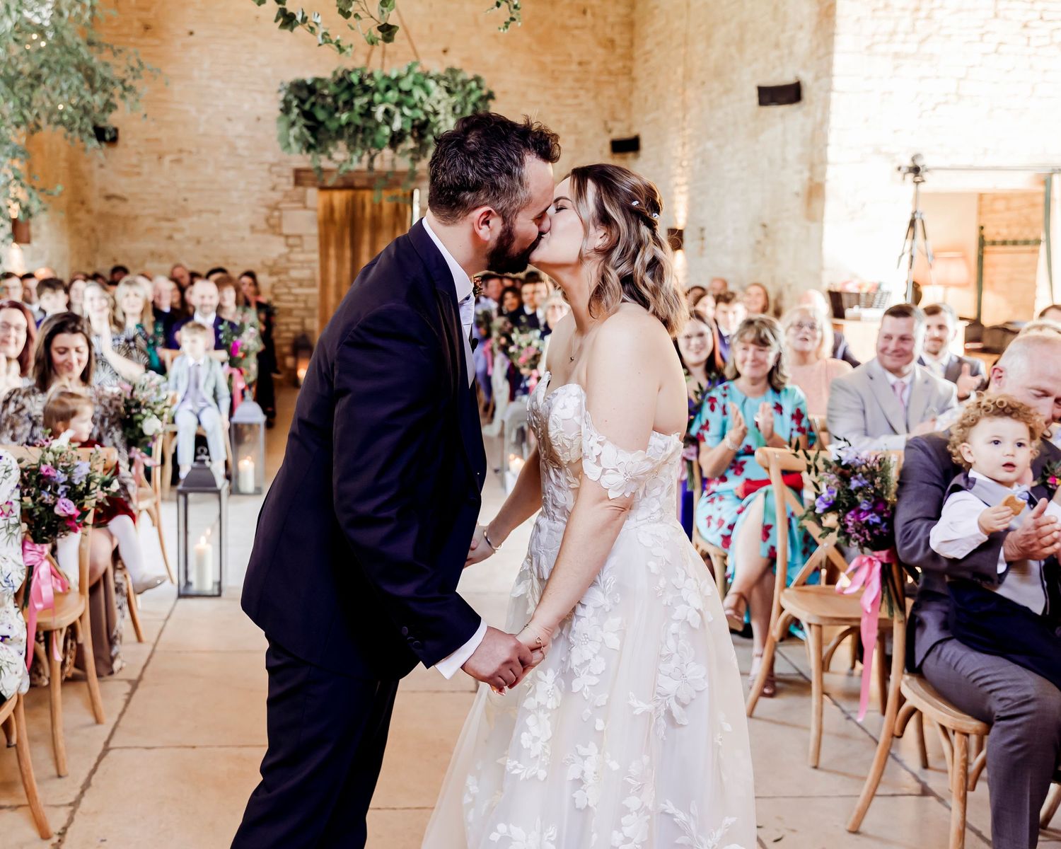 Bride and groom share first kiss at wedding ceremony with guests cheering in background.