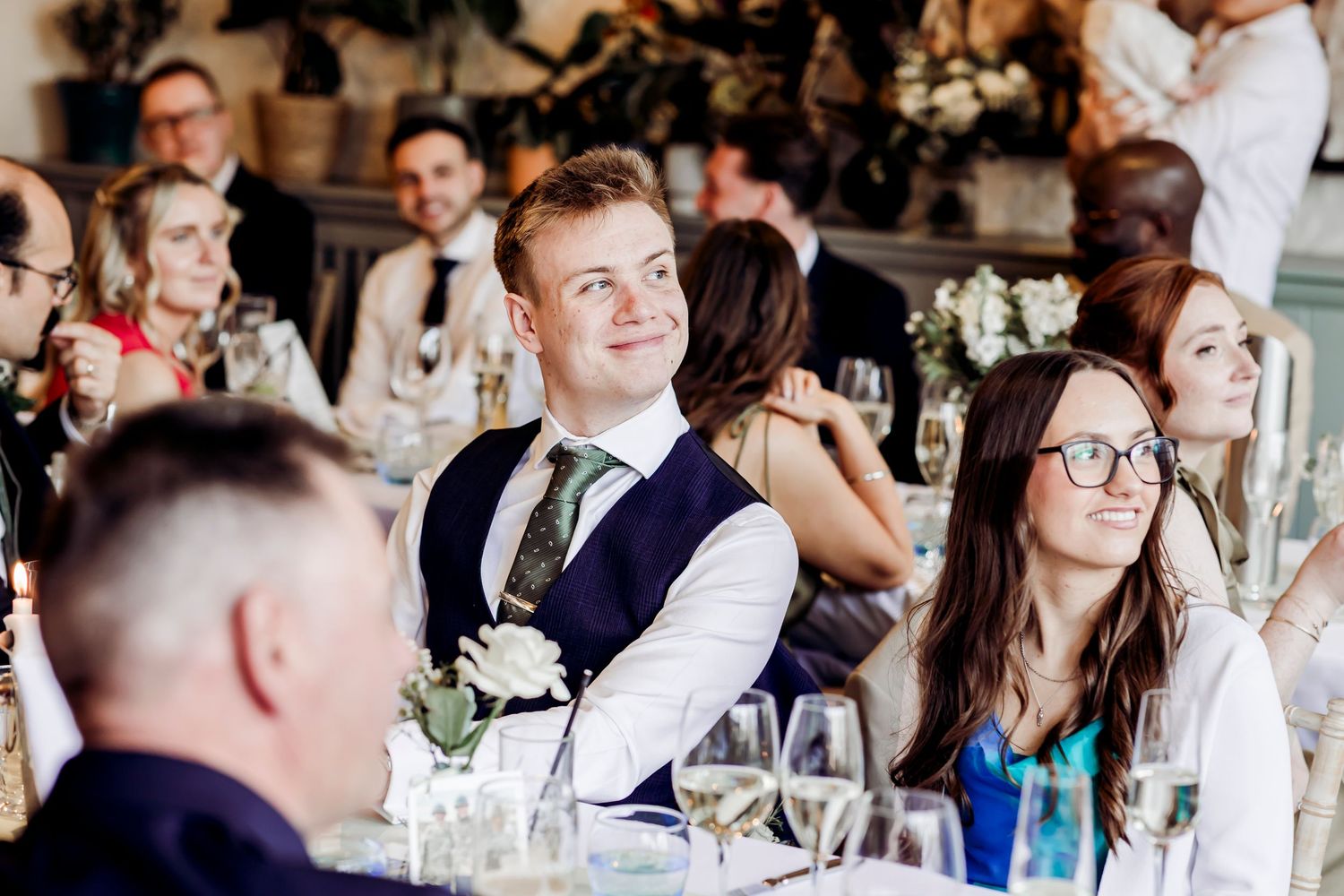 Wedding guests smiling and laughing during speeches at a bright reception venue.