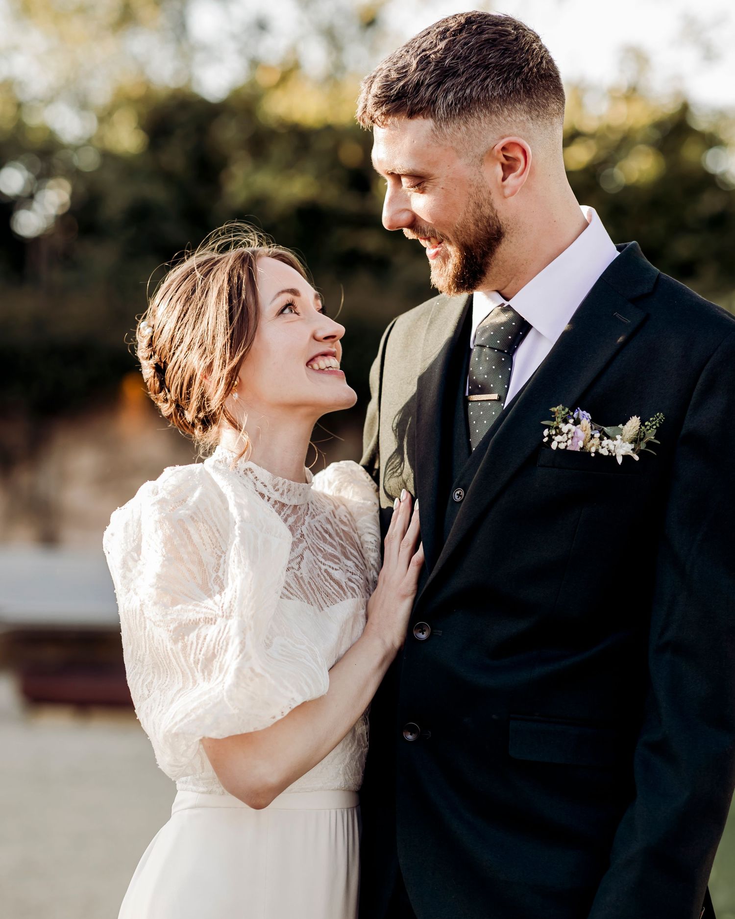 Smiling bride in vintage lace dress gazes lovingly at bearded groom in black suit at outdoor wedding.