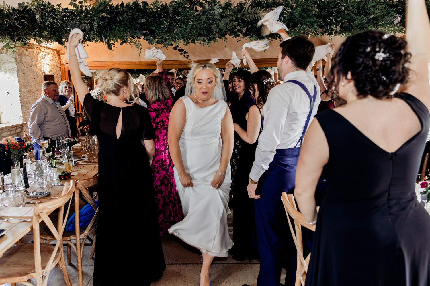 Bride in white satin gown dancing joyfully with wedding guests in a warmly lit rustic reception venue.