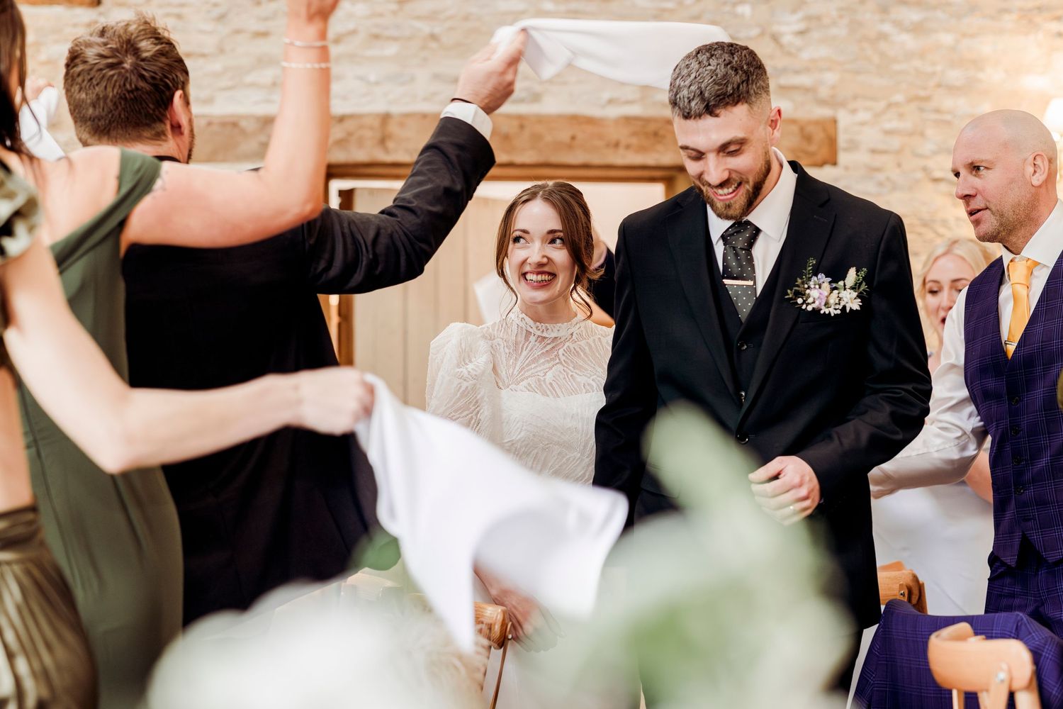 Joyful newlyweds walk through celebratory arch at rustic wedding reception venue.