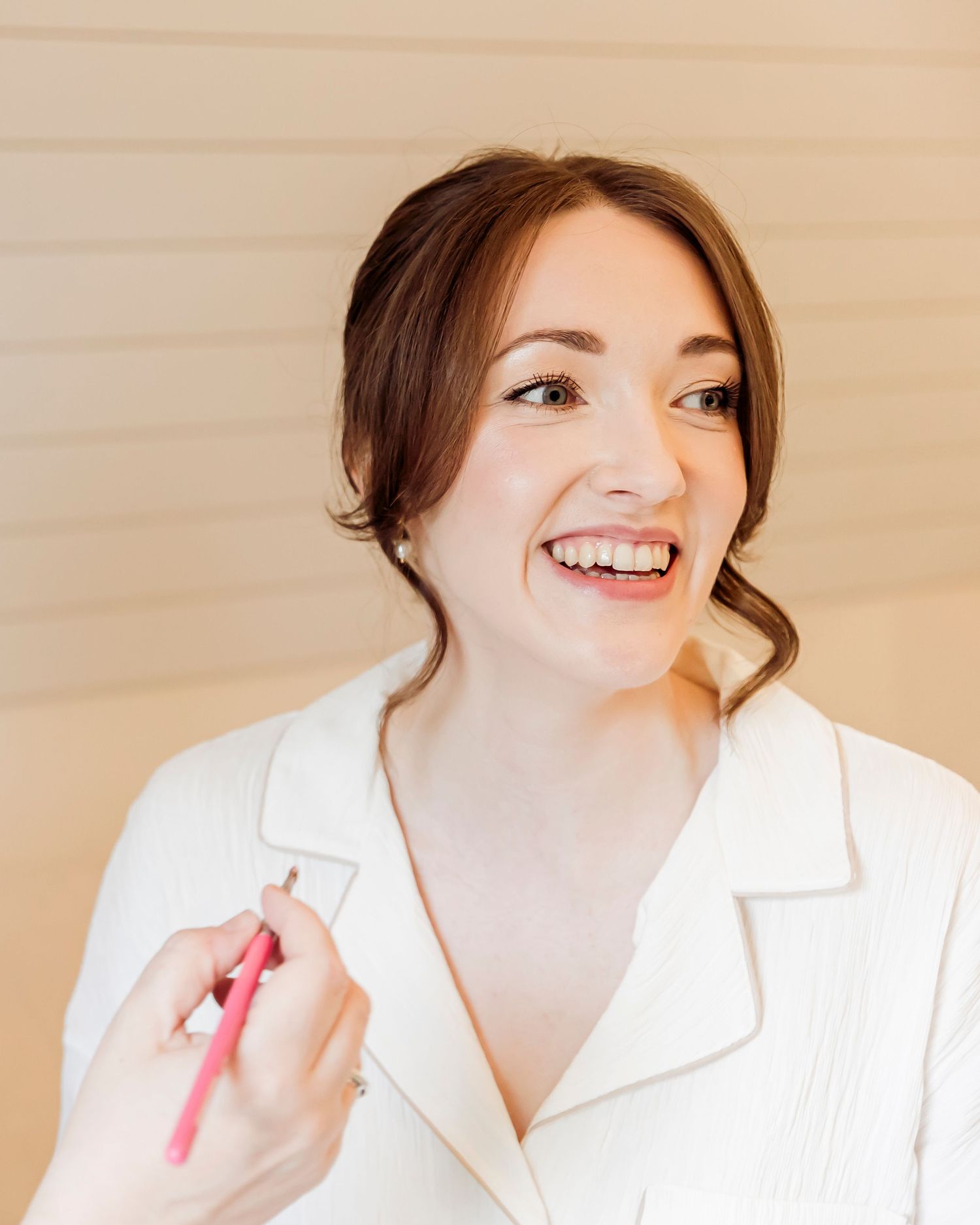 Smiling bride getting makeup applied with a pink brush, wearing a white robe during wedding preparations.