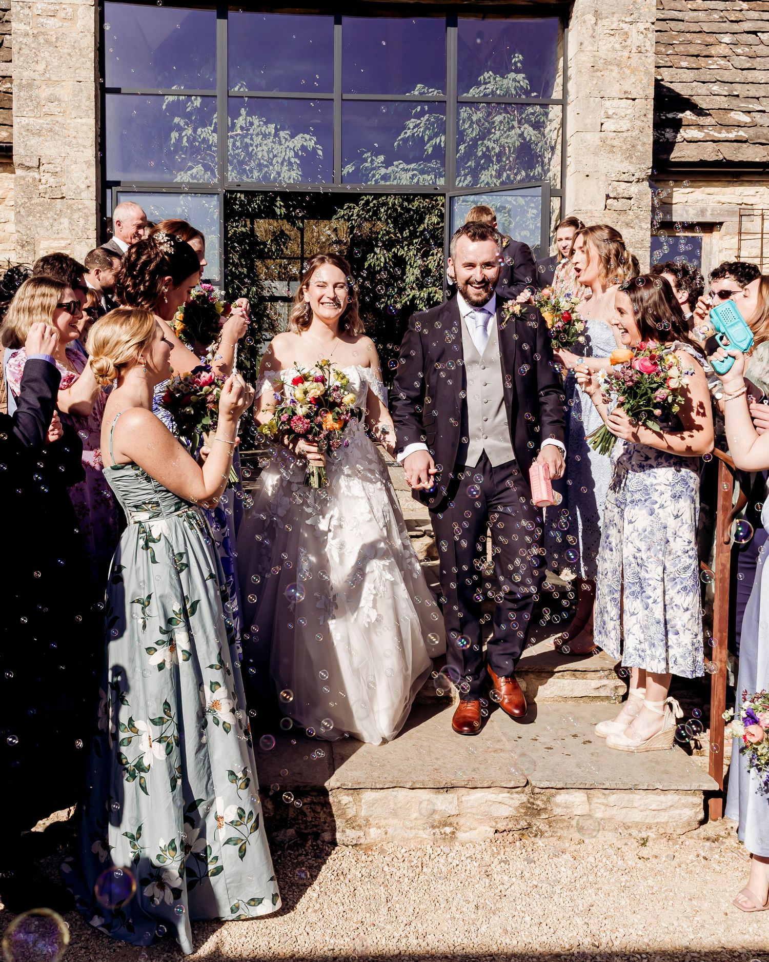 Bride and groom walk through confetti shower outside stone wedding venue with cheering guests.