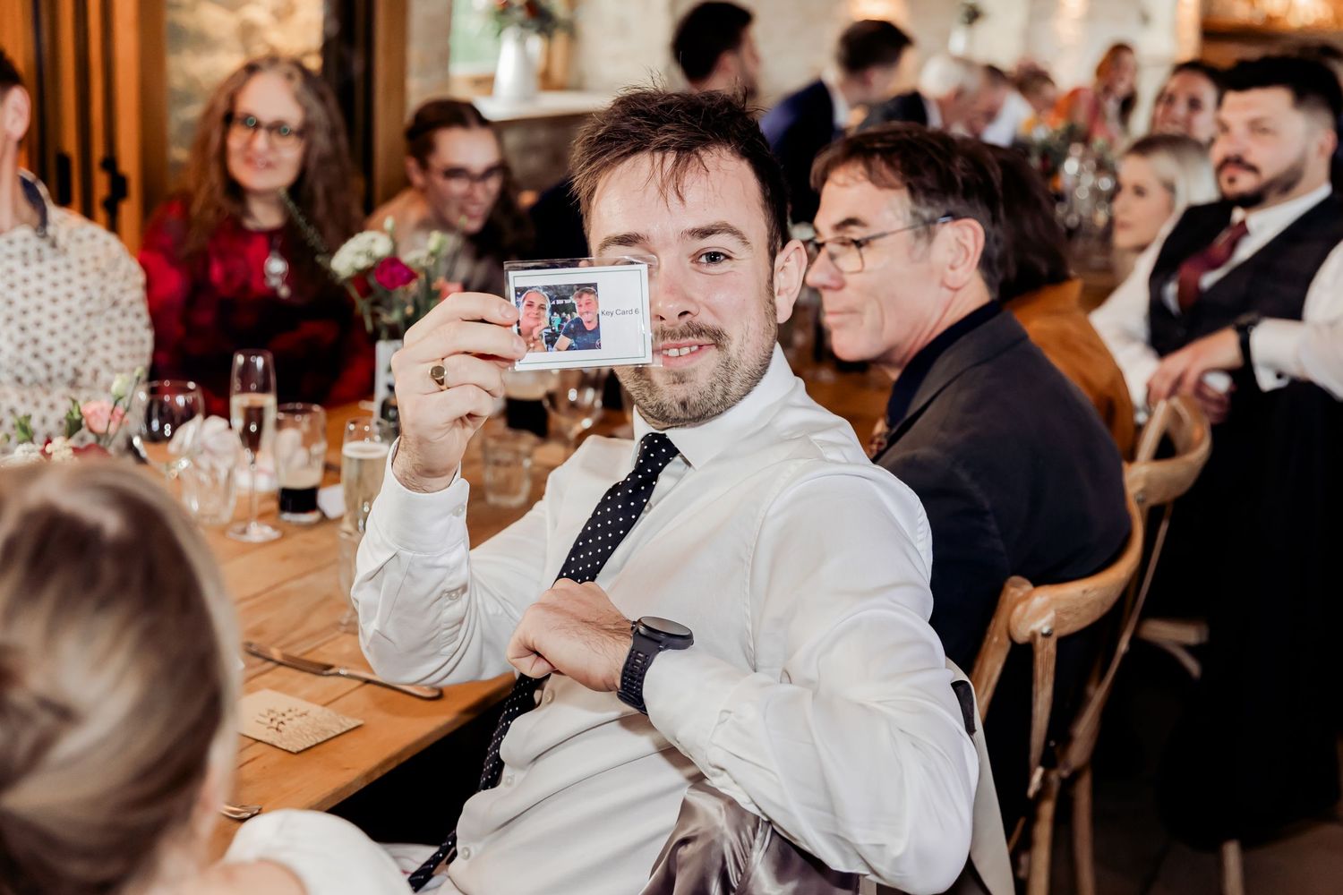 A wedding guest smiles while holding up a Polaroid photo at a rustic reception dinner.