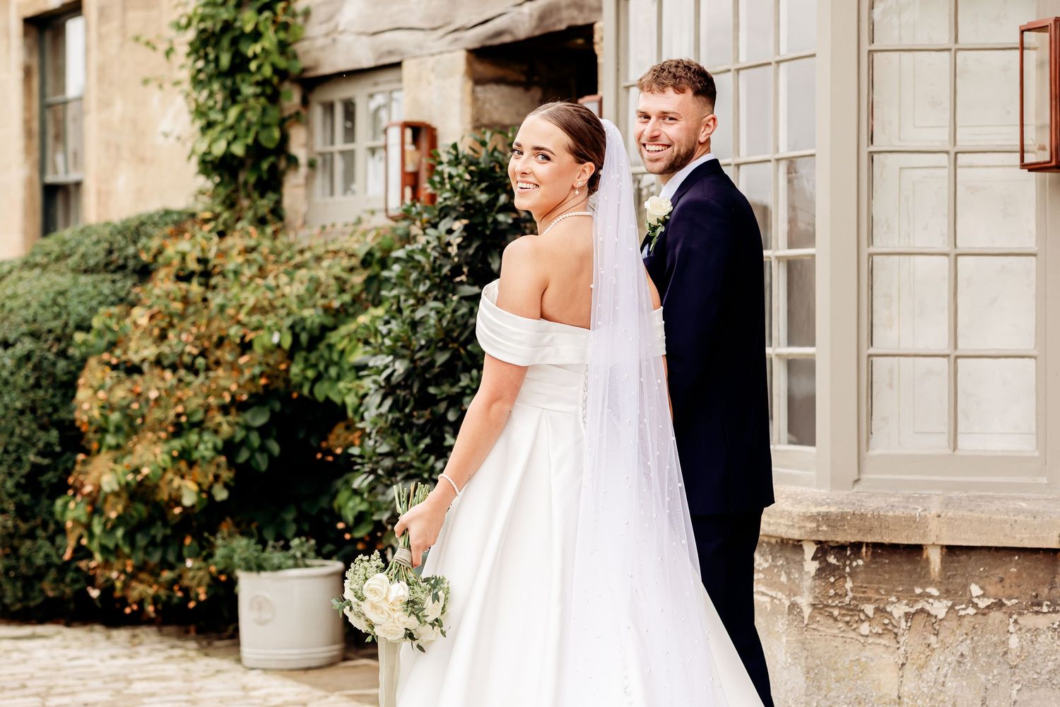 Elegant bride in off-shoulder gown holds white bouquet beside groom outside a classic stone wedding venue.