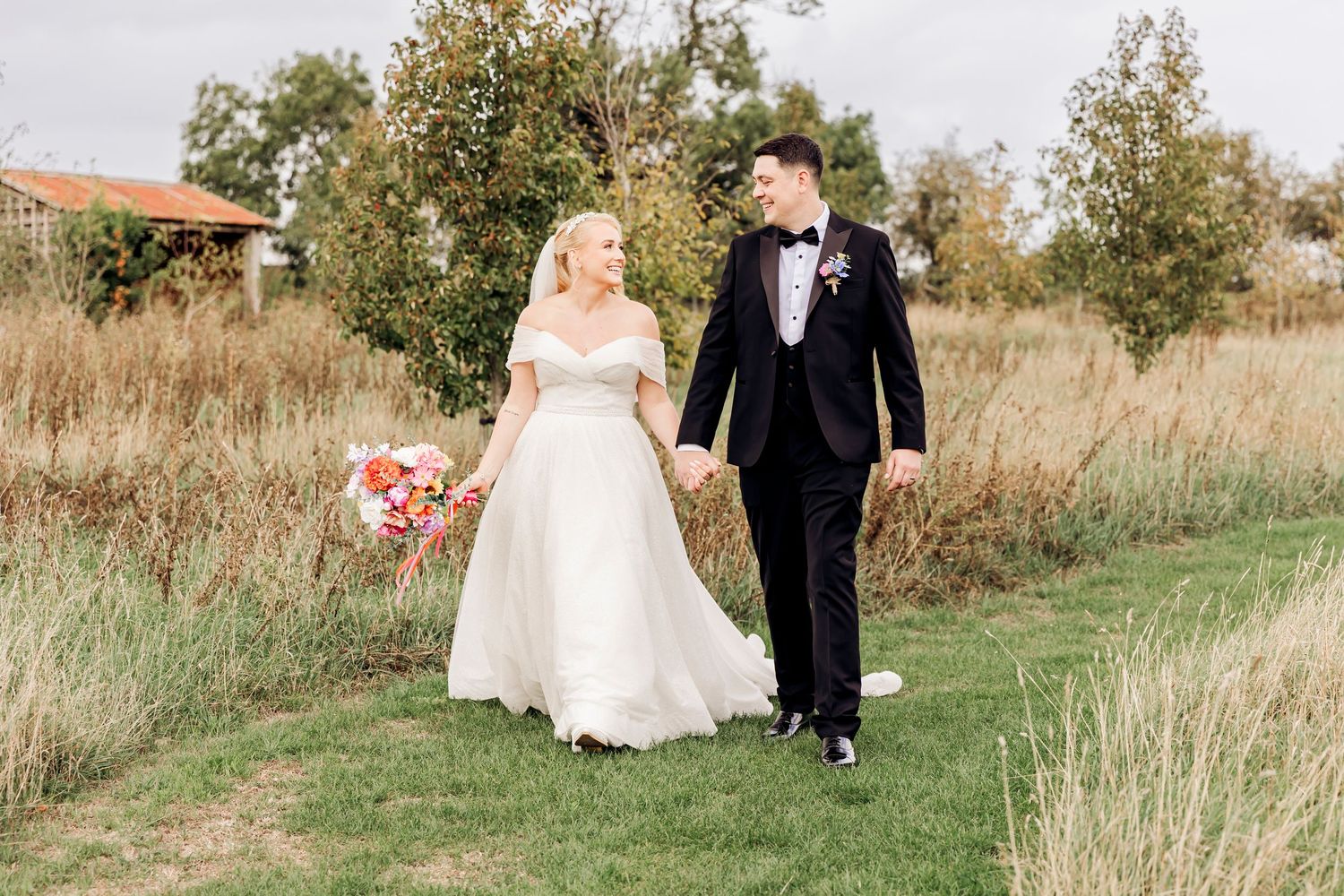 Bride and groom walk hand-in-hand through a lush green meadow path, bride holding a pink bouquet in wedding attire.