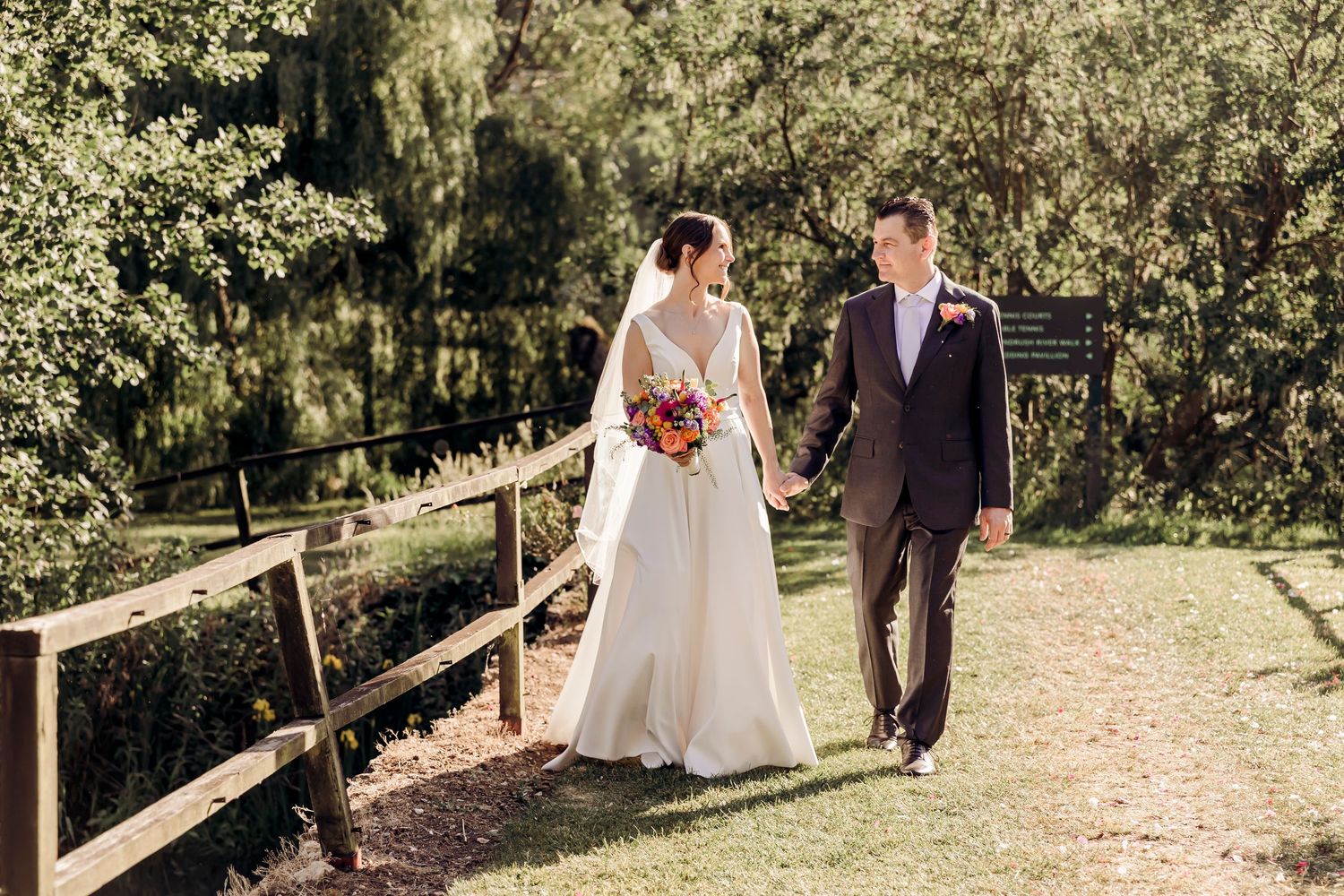 Bride and groom holding hands walking along a wooden fence path in a lush green garden setting.