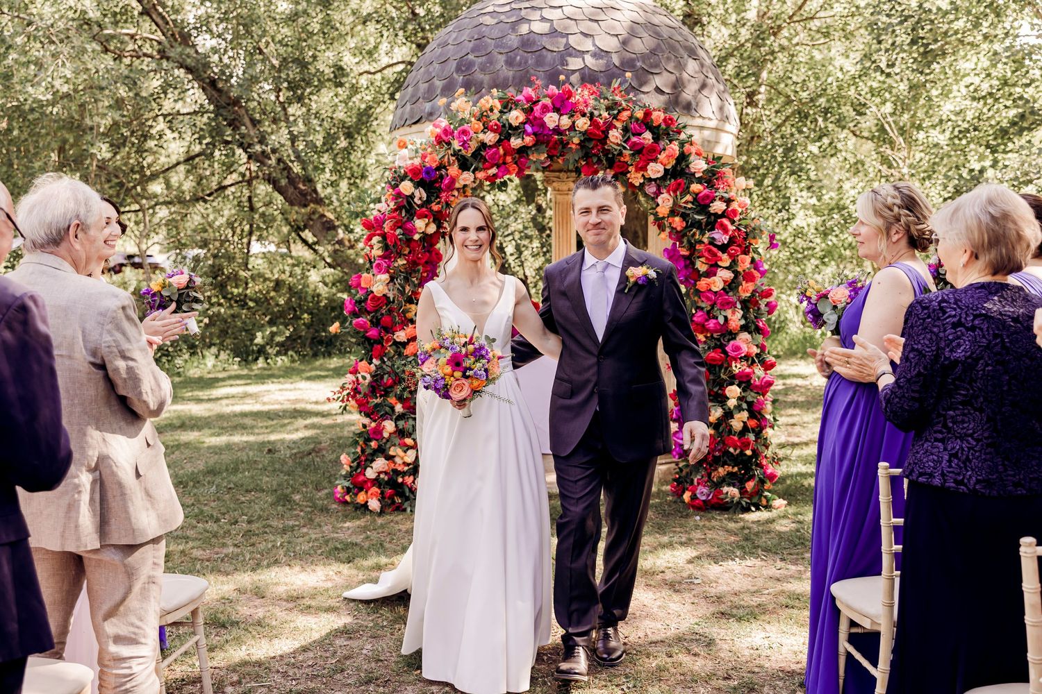 Newlyweds walk down aisle after outdoor wedding ceremony past vibrant pink floral arch with guests applauding in garden.