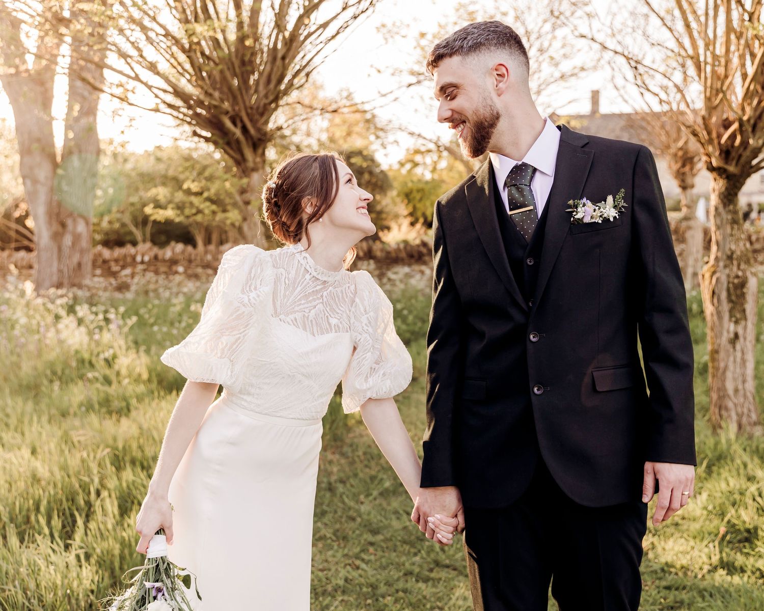 Newlywed couple holds hands and gazes at each other lovingly in a sunlit garden on their wedding day.
