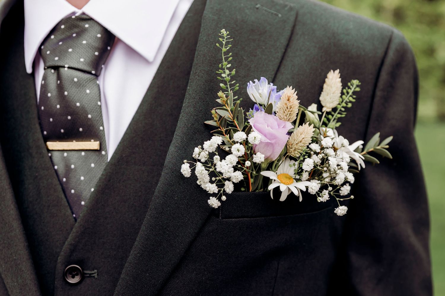 Close-up of a groom's dark suit with a floral boutonniere featuring purple, white, and wildflowers, and a polka dot tie.