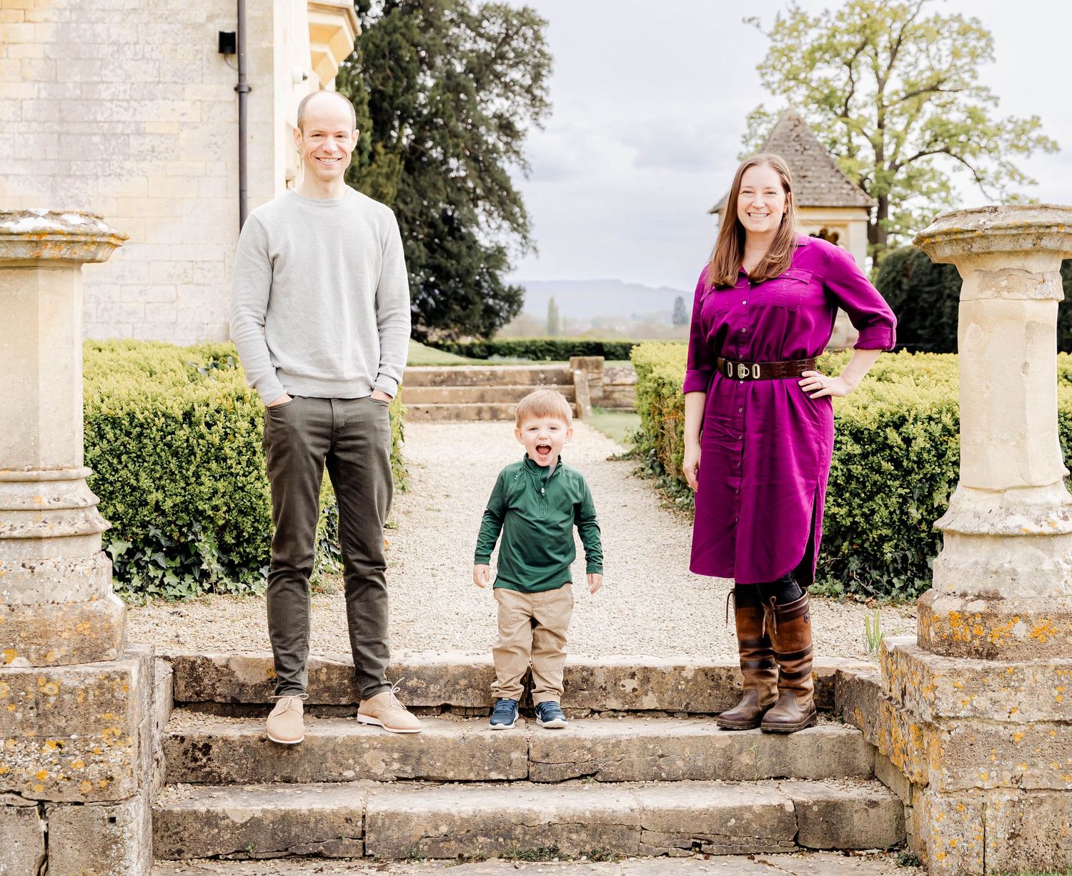 Family of three posing on stone steps outside a historic manor, parents flanking a young toddler boy.