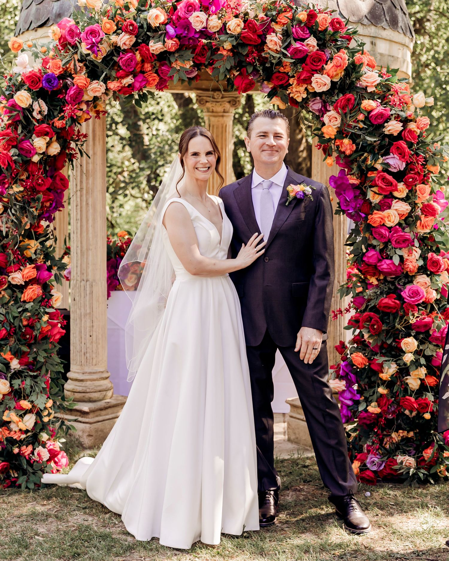 Bride and groom smiling under a vibrant floral arch of pink, orange, and red roses at an outdoor wedding ceremony.
