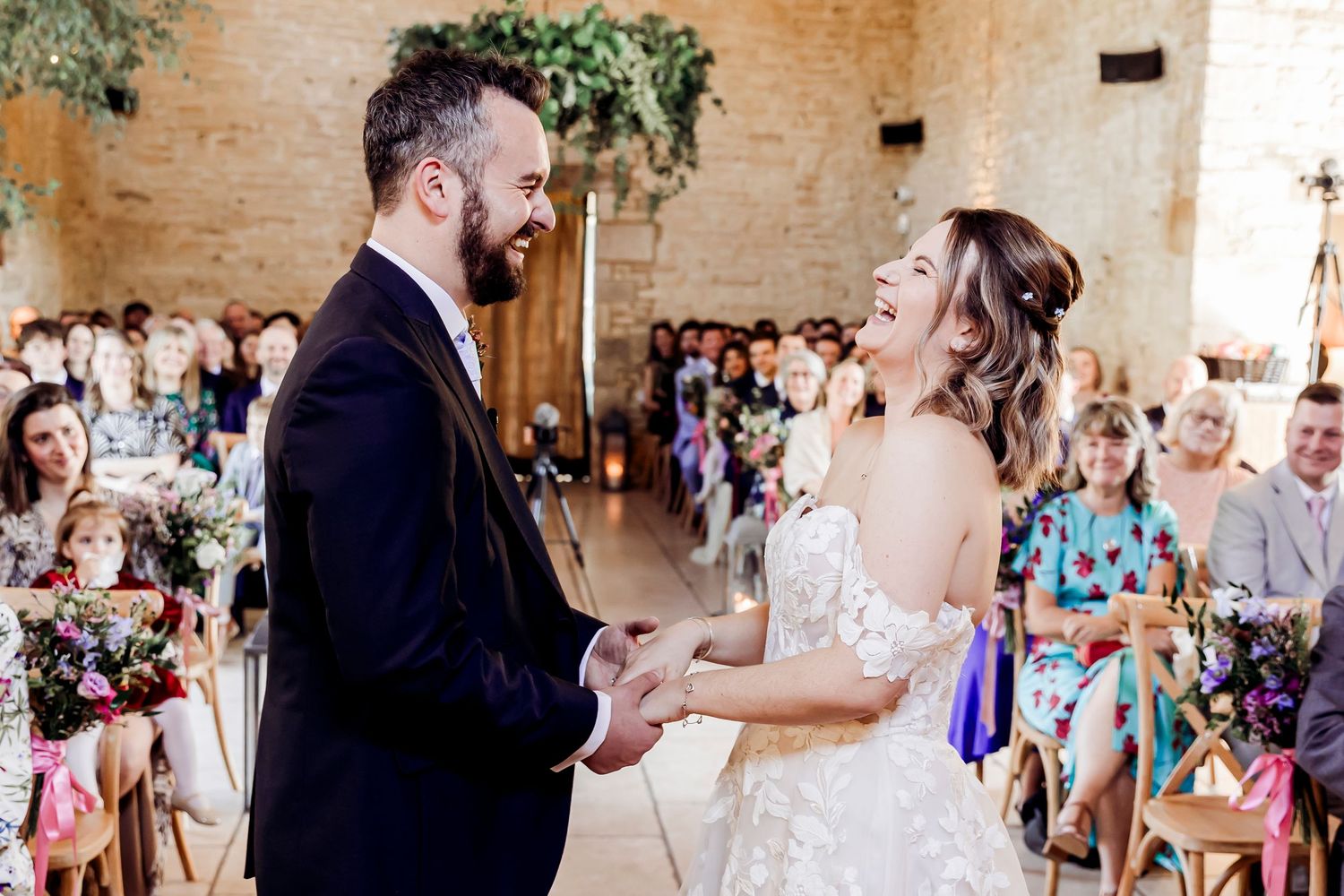 Joyful bride and groom laugh together holding hands during wedding ceremony in stone venue.