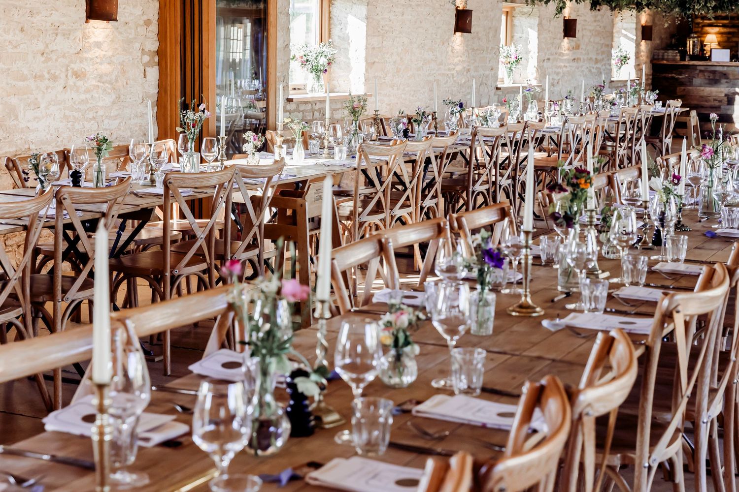Elegant rustic wedding reception hall with long wooden tables, cross-back chairs, floral centerpieces and candlesticks.