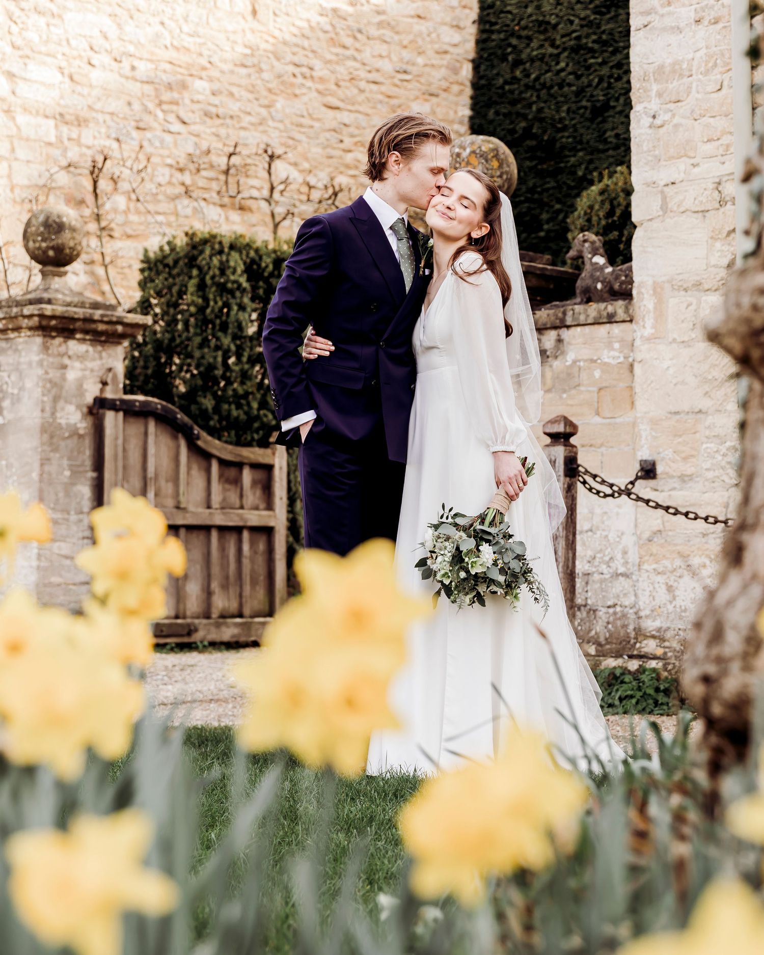 Wedding couple embracing near stone building surrounded by blooming yellow daffodils.