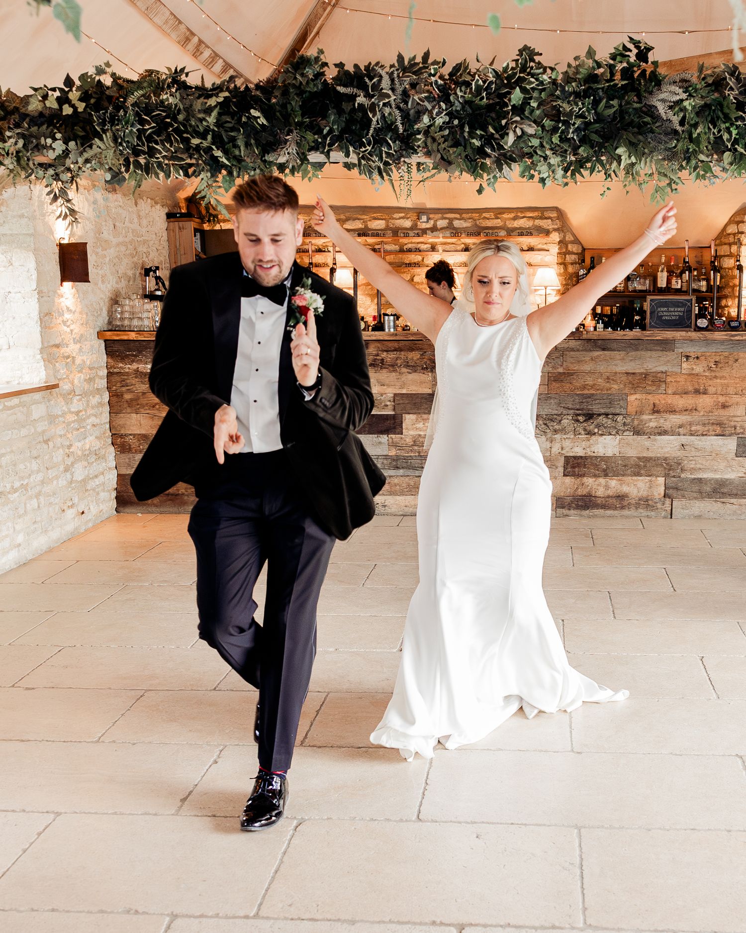 Excited bride and groom making grand entrance into rustic barn reception with arms raised in celebration.