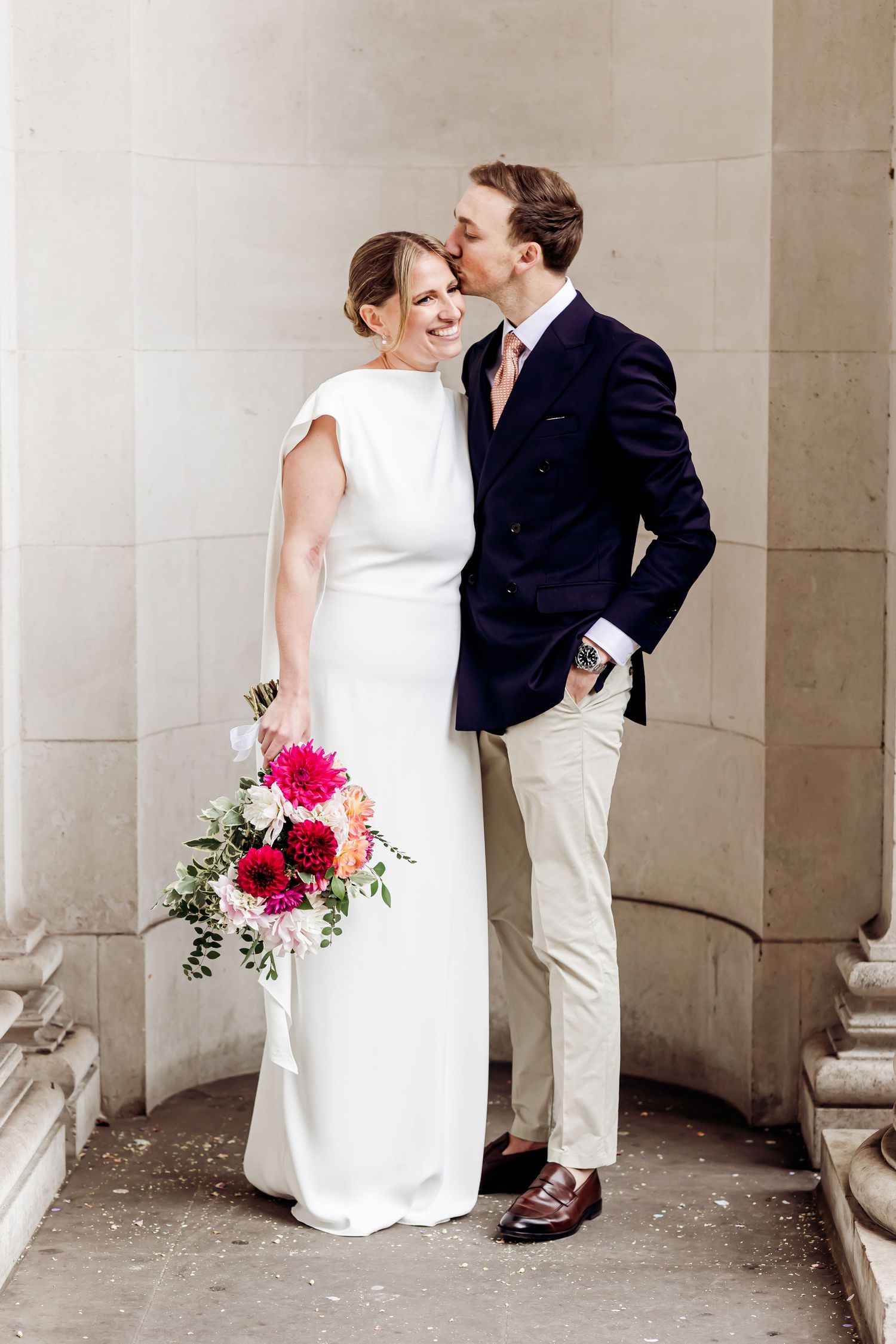 Groom in navy blazer kisses bride in sleek white gown holding vibrant pink bouquet at elegant venue.