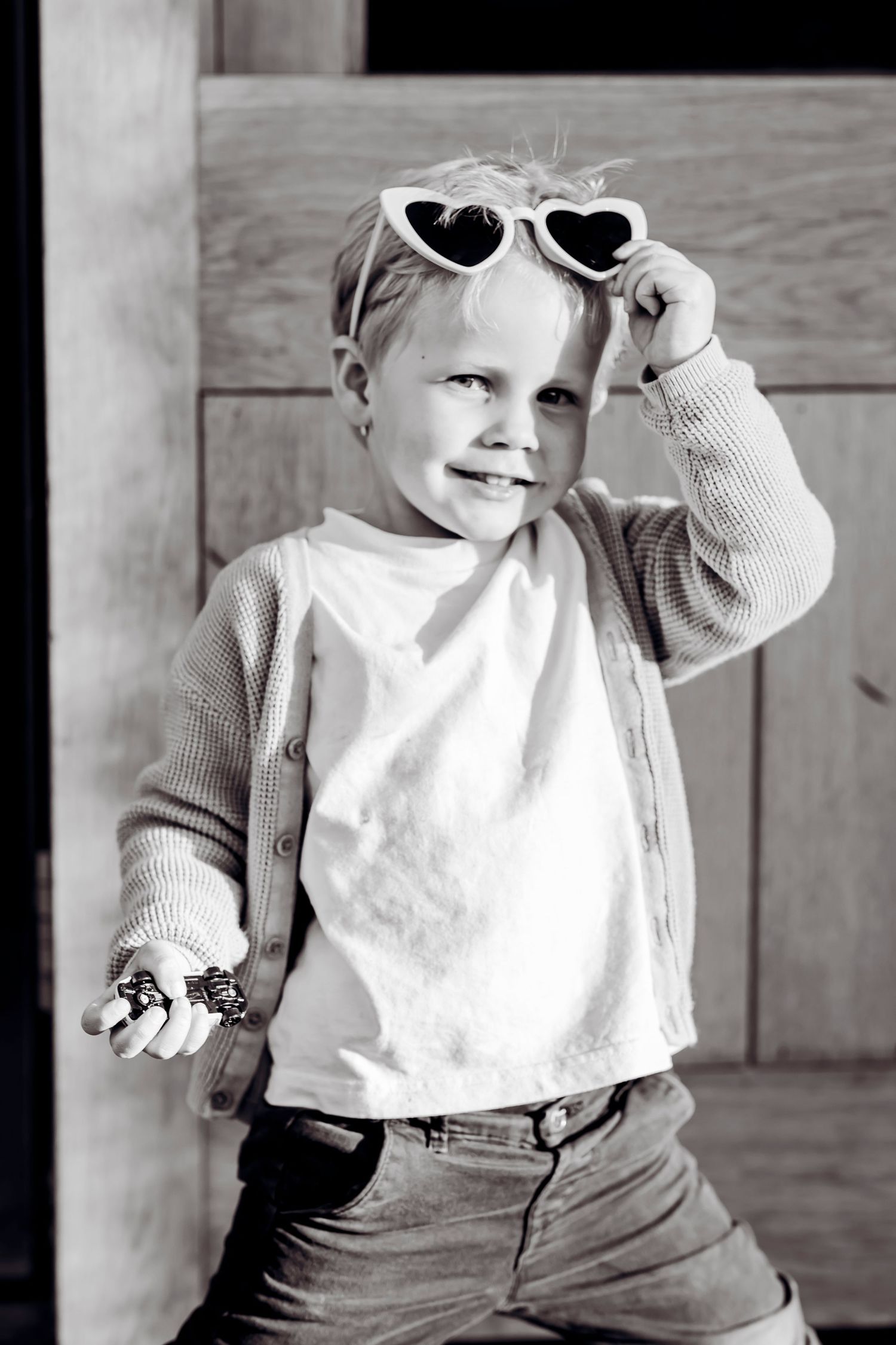 Cute young child in black and white photo wearing heart-shaped sunglasses and cardigan, smiling playfully.