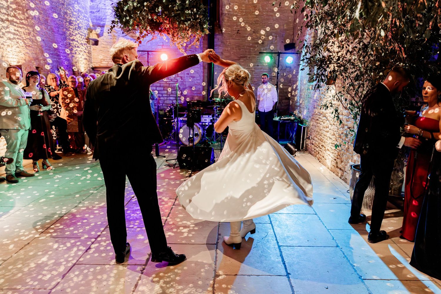 Groom twirls bride in flowing white wedding dress on dance floor with confetti and live band.