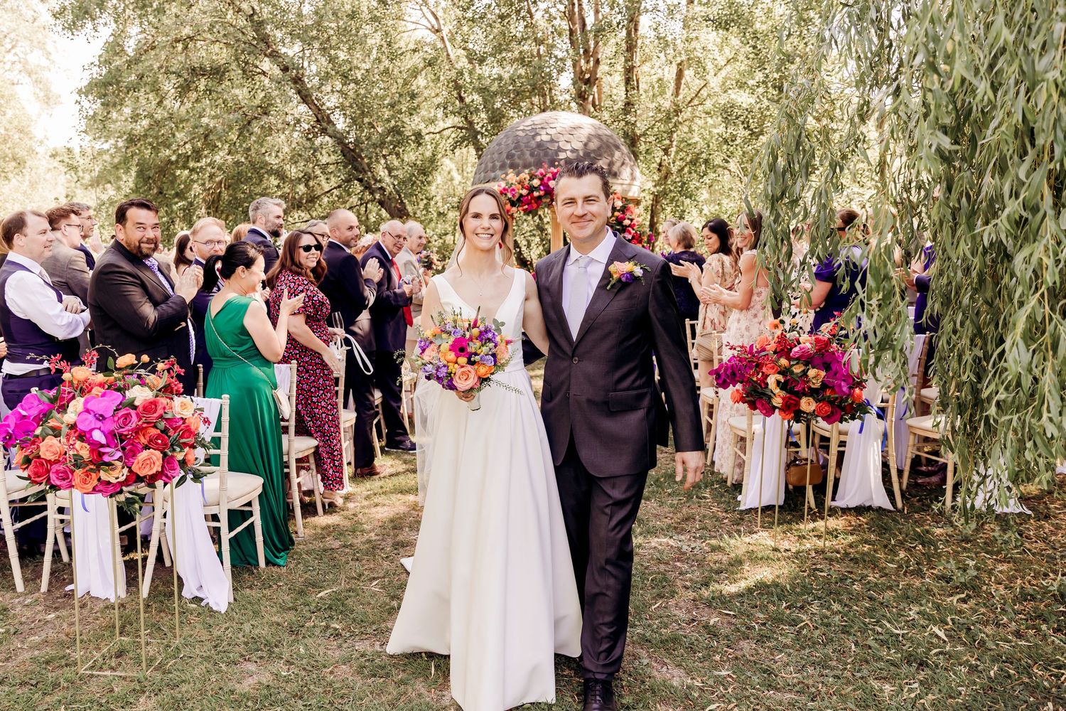 Bride and groom walk down outdoor aisle lined with vibrant pink and orange floral arrangements after wedding ceremony.