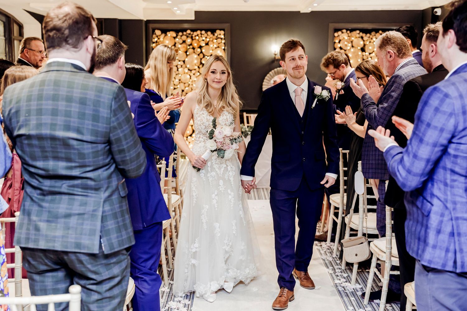 Newlyweds walking back down aisle as guests celebrate at elegant indoor wedding reception.