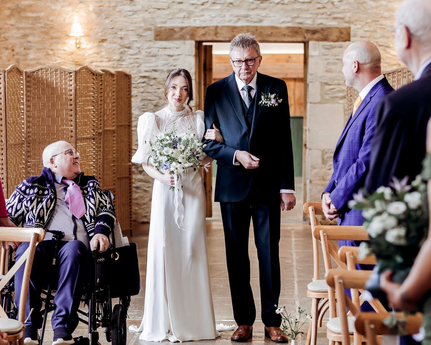Bride walking down the aisle with her father holding a bouquet in a rustic stone wedding venue.