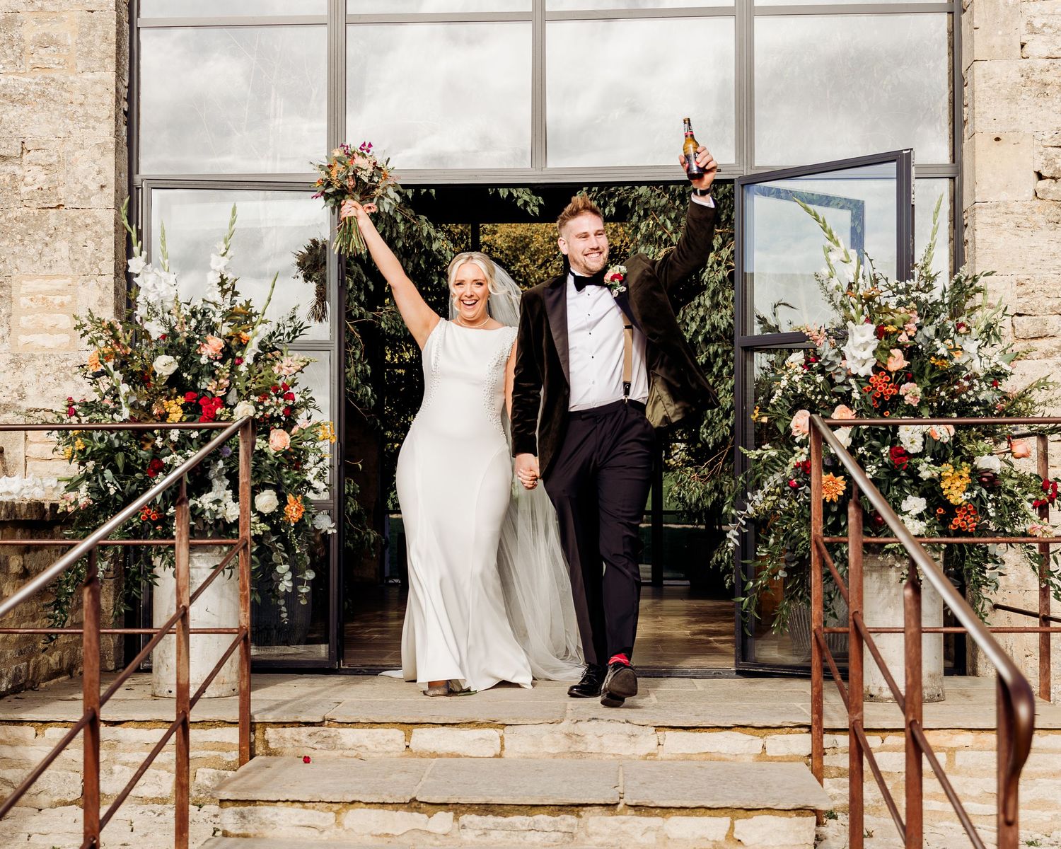 Joyful bride and groom celebrating at barn venue entrance, arms raised, surrounded by floral arch decorations.