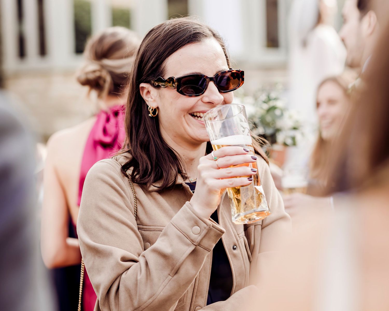 Woman in beige jacket and sunglasses smiling while drinking champagne at outdoor wedding.