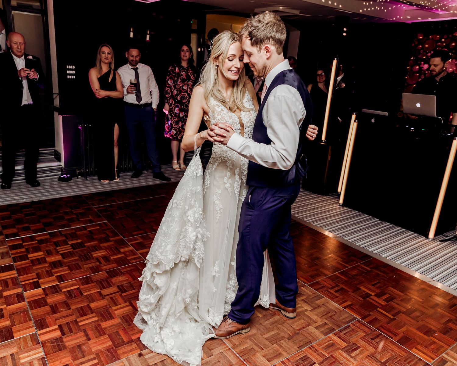 Bride in a floral lace gown and groom in a blue suit share a romantic first dance at their wedding reception.